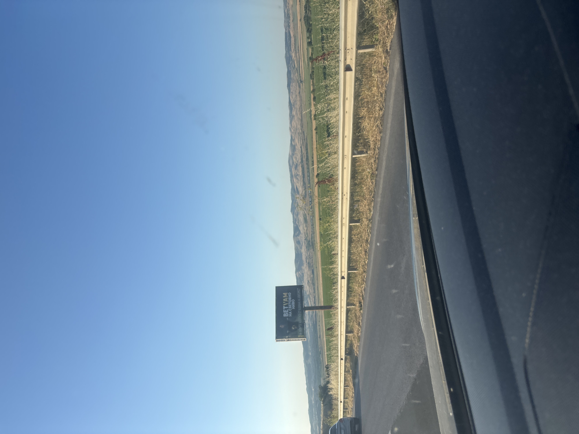 A billboard with the text "BETVAM НА СВЕТОВНО НИВО" is visible on the side of a highway under a clear blue sky. The billboard is positioned in front of a landscape of rolling hills and agricultural fields. A white metal guardrail runs along the side of the highway, separating it from the grassy verge and fields. A car is seen driving on the highway to the left of the frame. The foreground shows the dashboard of a vehicle.