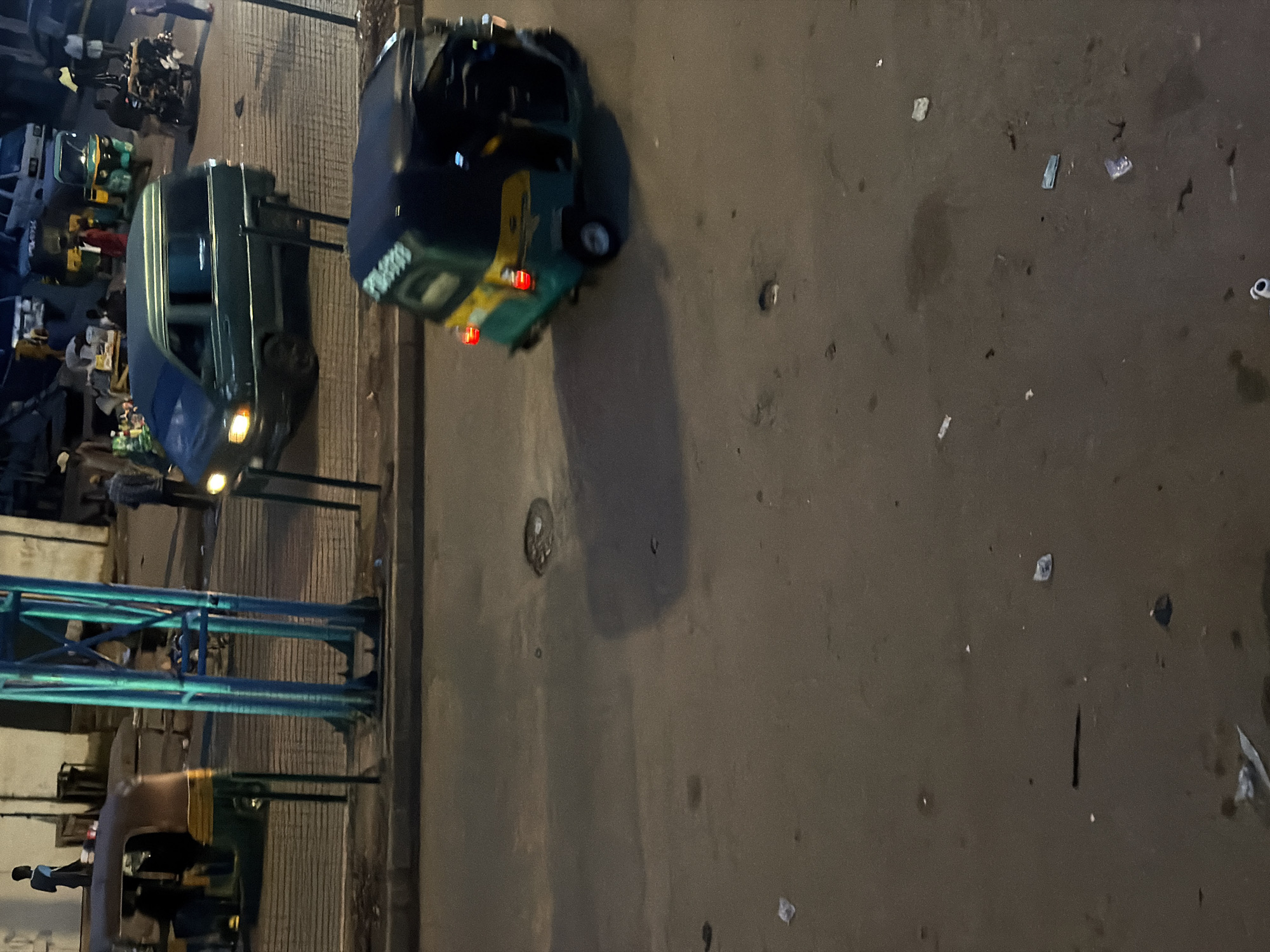 The image depicts a street scene at night in Bukuru, Nigeria, with a focus on vehicular and pedestrian activity. In the foreground, a green and yellow auto-rickshaw is captured in motion, its brake lights illuminated. The street surface appears wet and somewhat uneven, with scattered debris. In the mid-ground, a dark green van is parked on a paved area, possibly a sidewalk or market stall area. A teal blue traffic light structure is prominent on the left side of the frame, its lights not discernibly on. Beyond the van, several people are visible, some standing and interacting, while others appear to be moving. Further in the background, a white vehicle and another auto-rickshaw can be seen, suggesting a busy urban environment. The lighting is dim, characteristic of nighttime conditions, with artificial lights from vehicles and possibly streetlamps creating shadows and highlights. The overall atmosphere suggests a typical evening in a bustling Nigerian city.