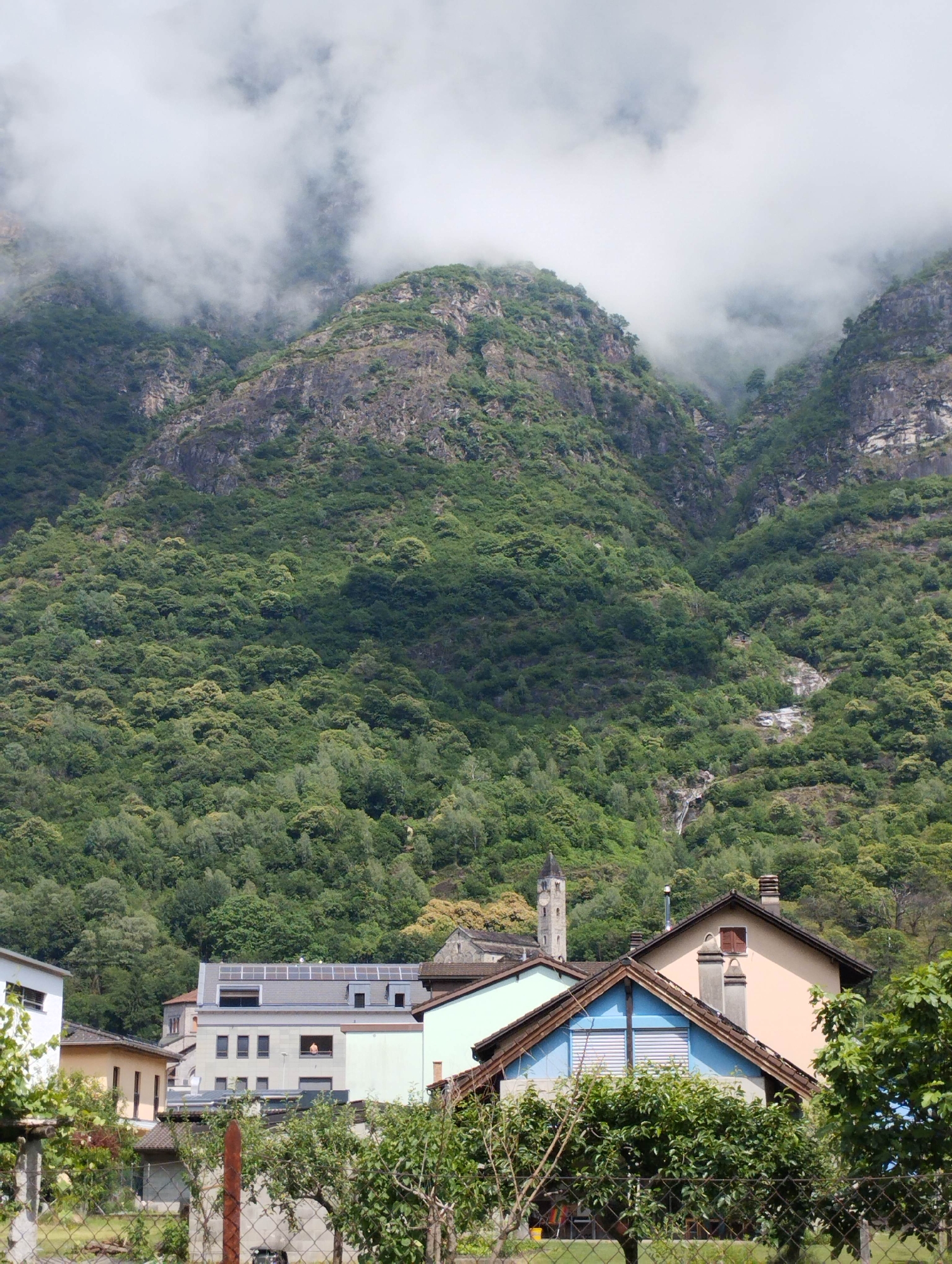The image displays a panoramic view of a village situated at the base of large, steep mountains, under an overcast sky. The upper and mid-sections of the mountains are shrouded in thick, white and grey clouds or mist, which partially obscure the peaks. The mountains are densely covered with varying shades of green foliage and trees, interspersed with visible rocky outcroppings and cliffs. A thin stream or waterfall can be seen descending a distant part of the mountain on the right side.
In the midground, a cluster of residential buildings with diverse architectural styles and roof colors is visible. One prominent building features a dark grey roof equipped with multiple solar panels. A single person, appearing to be an adult, is discernible within an open window or balcony of this building, looking outwards. Closer to the foreground, a house with a peach-colored facade and multiple chimneys stands adjacent to another house with a distinctive blue-painted upper gable fitted with white horizontal blinds. A tall, slender church tower, featuring a pointed roof and visible clock faces, rises above the other village structures, positioned towards the left of the central cluster of buildings.
The immediate foreground includes a wire mesh fence running horizontally across the bottom of the image, behind which are various green shrubs and trees, some appearing to be fruit-bearing. The overall scene is characterized by lush greenery and a natural setting, with the village nestled within a dramatic mountainous landscape in the city of Unknown, Unknown.
