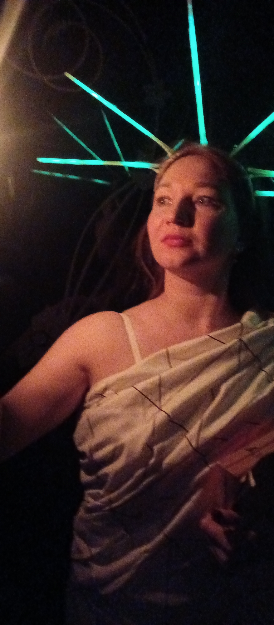 A woman is standing in a dark environment with green neon light fixtures overhead. She is wearing a light-colored dress with thin straps. The location is Unknown, Unknown.