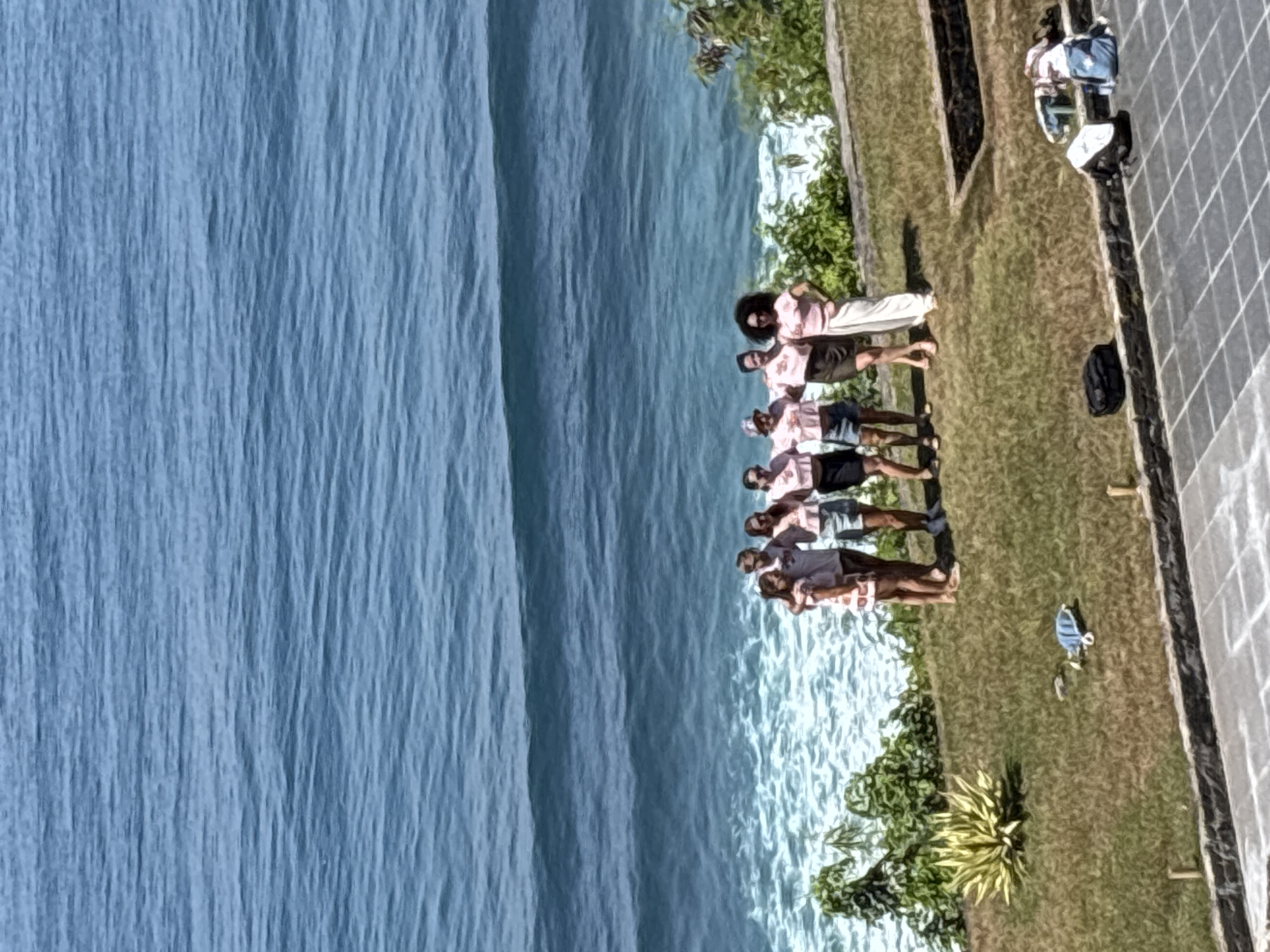 The image shows a group of approximately 7 people standing on a grassy area near the ocean in Kuta Selatan, Indonesia. They are positioned near a coastline with trees and greenery. The people appear to be wearing similar pink shirts. Adjacent to the grassy area is a tiled or paved surface. Beyond the grassy area, the ocean is visible.