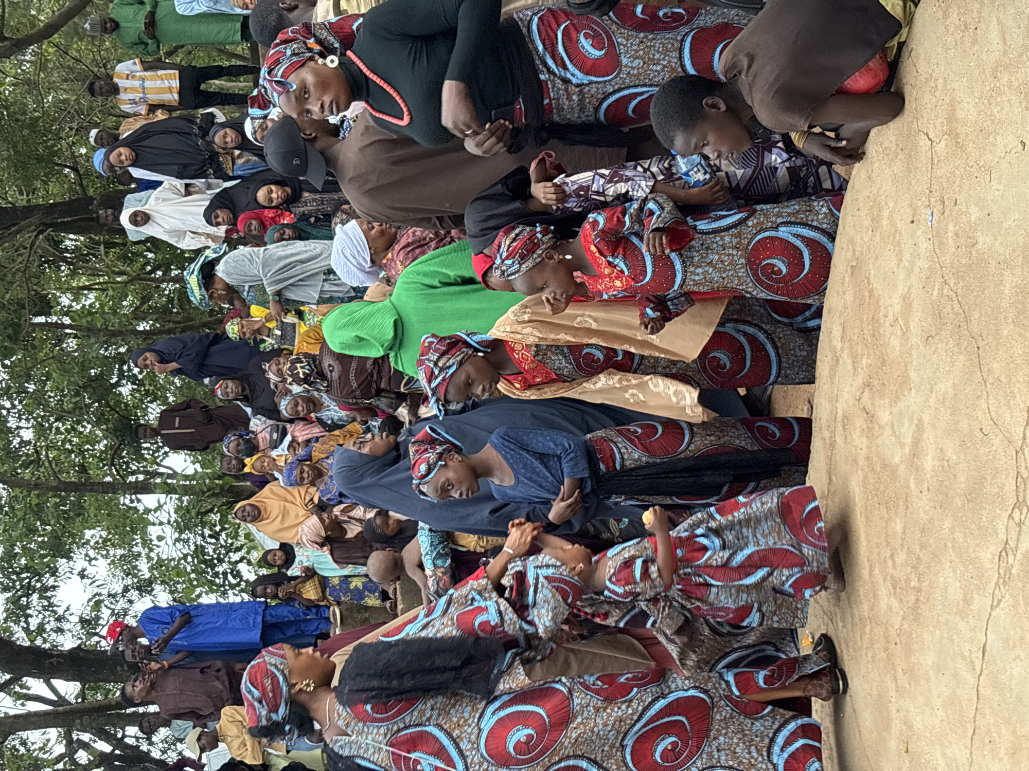 The image depicts a gathering of people, predominantly women and children, in what appears to be an outdoor event. The individuals are dressed in colorful, patterned traditional attire, featuring vibrant reds, blues, and browns. Many women are wearing headscarves, and the children are also adorned in similar festive clothing. The setting suggests a community event or celebration, possibly a religious or cultural ceremony. The background shows trees, indicating an outdoor location. While specific landmarks are not visible, the presence of a diverse group in traditional African clothing could be consistent with various cultural events in Nigeria. The individuals are standing and some are interacting, with children at the forefront appearing engaged in the event. The lighting suggests daylight, possibly overcast.