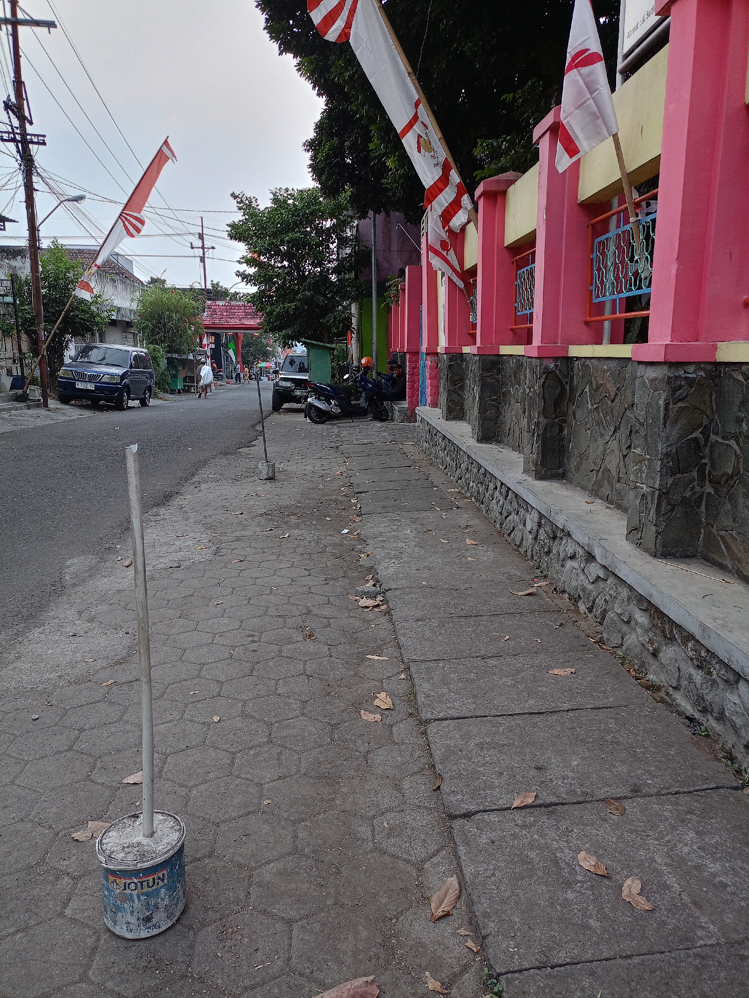 The image depicts a street scene in Batu, Indonesia, featuring a sidewalk, a road, and buildings. The sidewalk is paved with hexagonal tiles and concrete slabs, with scattered dry leaves. A white pole is planted in a blue Jotun paint bucket filled with concrete, positioned on the sidewalk.
The road is asphalted and appears to be a two-lane street. Several vehicles are present: a dark blue SUV is parked on the left side of the road, and a motorcycle is parked on the right side. Further down the road, another vehicle, possibly a van, is visible.
Along the right side of the sidewalk, a pink building with yellow accents and a stone-clad foundation is prominent. Red and white flags, possibly national flags, are displayed on poles along the top of the pink building's facade. The flags are horizontal, with a red stripe at the top and a white stripe at the bottom. Some flags have additional ornamentation. Trees with dense green foliage are situated behind the flags and the pink building.
In the background, electric poles with numerous wires are visible, indicating the presence of urban infrastructure. A few people are walking on the sidewalk and the road in the distance, suggesting a public area with some activity. The sky is overcast, creating a diffused light. The overall atmosphere suggests a typical street environment in a populated area.