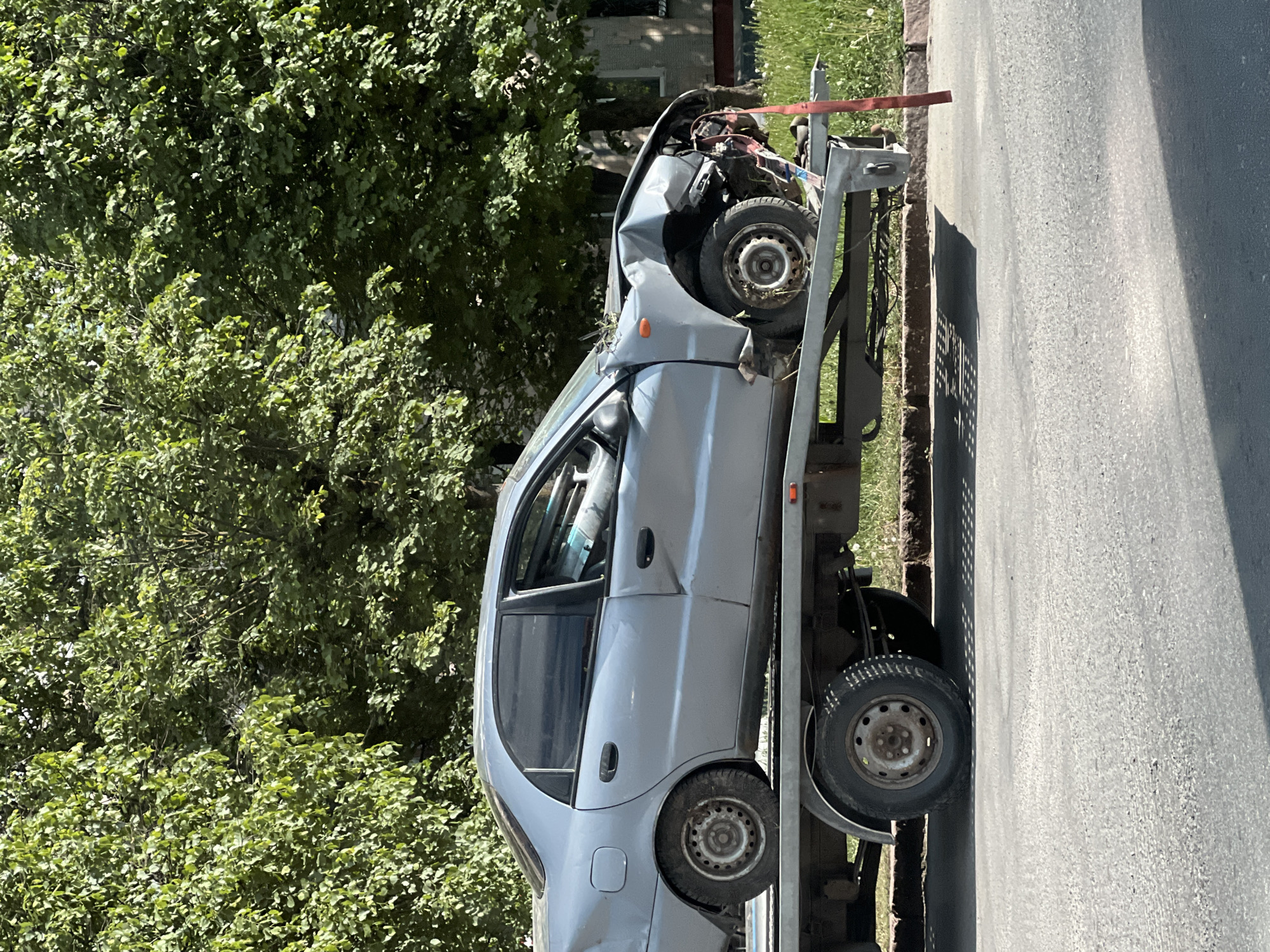 The image shows a severely damaged light gray car on a tow truck. The front of the car is heavily crushed, indicating a major impact. The tow truck is parked on a paved road, with green trees and foliage visible in the background. Based on the context provided, the location is likely in the city of Ternopil, Ukraine. There are no people visible in the image. The overall scene suggests the aftermath of a significant car accident.