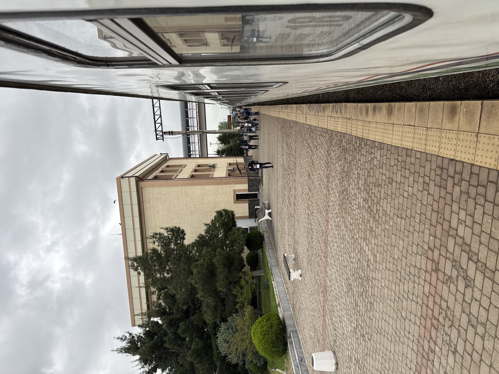 The image depicts a train station platform in Jizzakh, Uzbekistan, during overcast weather. A train is parallel to the platform on the right. A group of people is congregated further down the platform, possibly waiting for or disembarking from the train. The platform is constructed of red and gray pavers with a yellow safety strip near the train tracks. Adjacent to the platform is a landscaped area with trees and benches. A multi-story building is present, partially visible behind the trees.