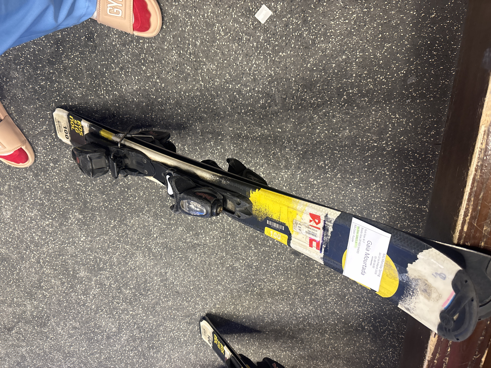The image shows a pair of skis with bindings lying on a speckled grey floor, possibly in a locker room or storage area. The skis are dark blue with yellow and white accents. One ski has a label that reads "Gaia Mourinho" and appears to have information about ski size, boot size, and color. To the right of the skis, a person wearing blue trousers and light-colored slides with red inserts is partially visible. Another person's legs and feet, wearing similar slides, are visible in the upper left corner of the image. The environment appears to be indoors, with a wooden bench or ledge in the foreground. There is no clear indication of the specific location within Dubai, United Arab Emirates.