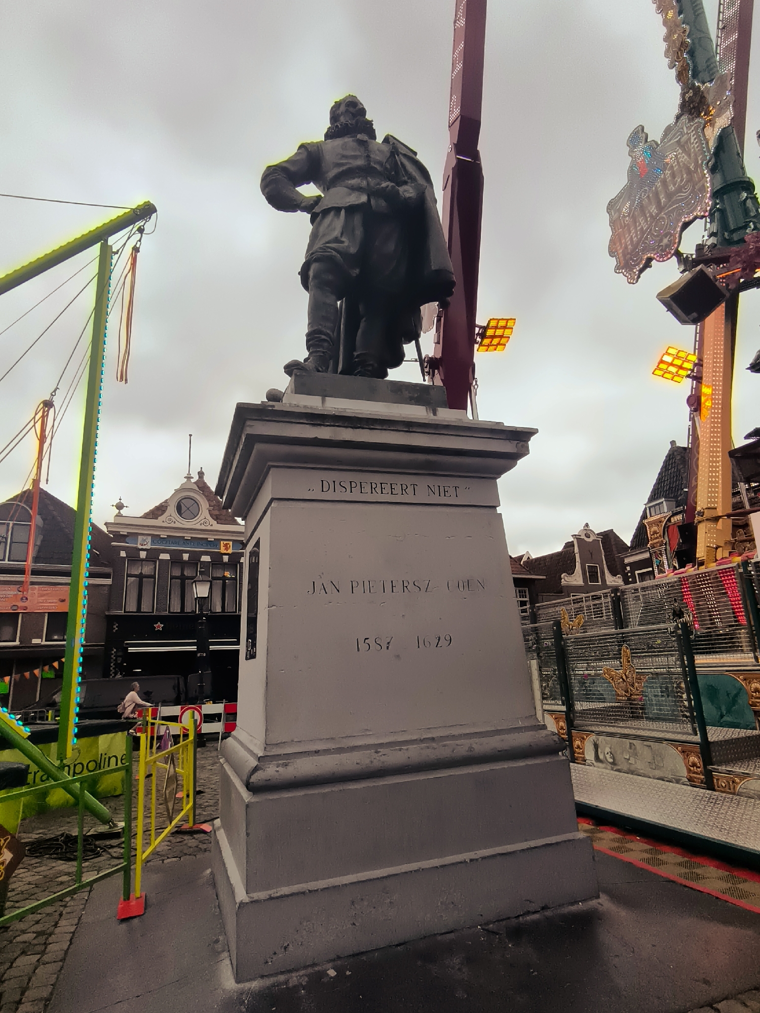 The image shows a statue of Jan Pieterszoon Coen in the city of Hoorn, Netherlands, on a cloudy day. The statue stands on a pedestal with inscriptions. Behind the statue, there are buildings with characteristic Dutch architecture, and carnival rides with colorful lights are visible. A trampoline and a yellow safety fence are in the foreground. The scene appears to be a public space, possibly a square or street, with a mix of historical elements and modern entertainment attractions.