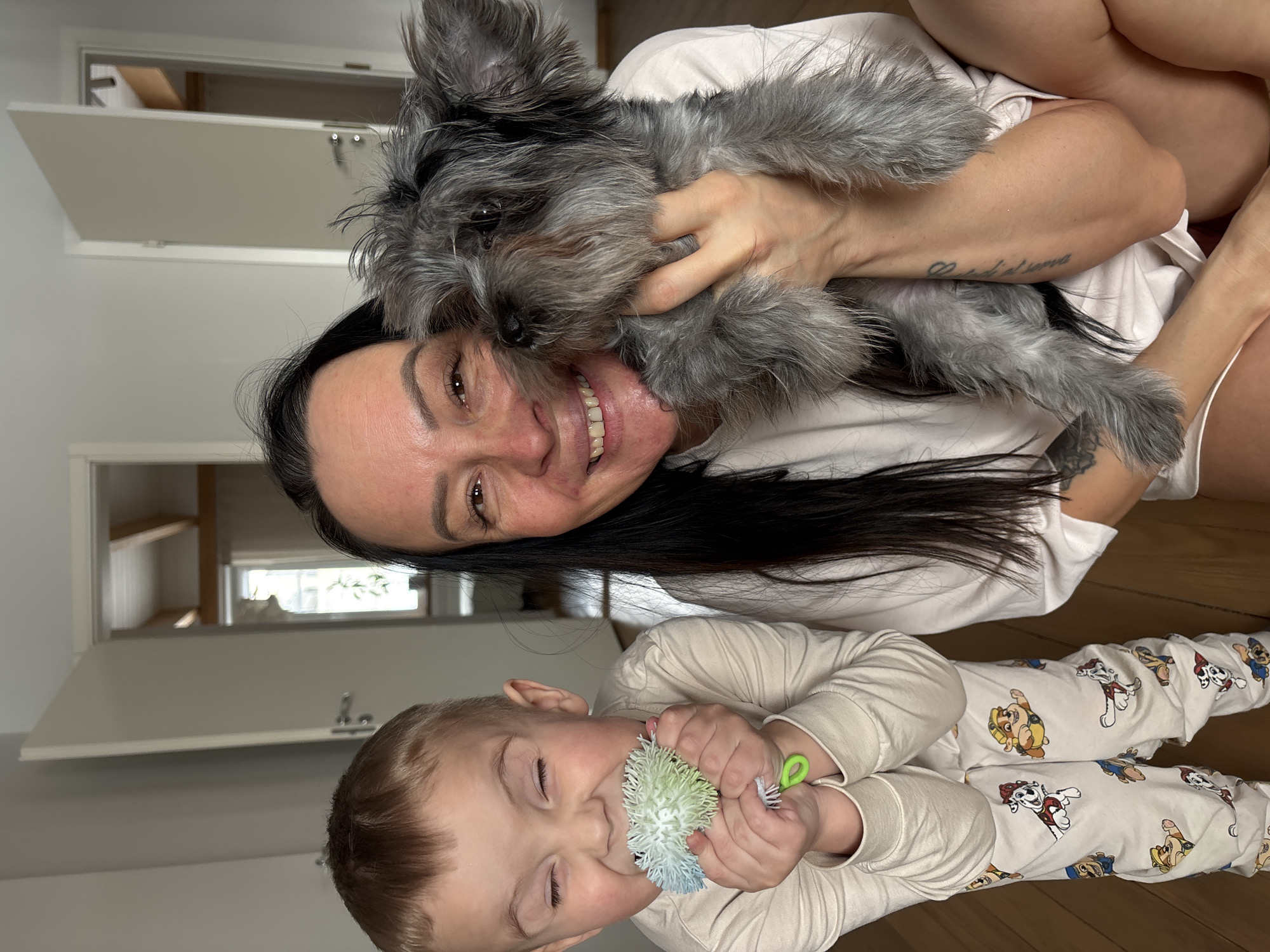 A photograph captures a woman, a child, and a dog in what appears to be an indoor setting. The woman, with dark hair and visible tattoo on her arm, is holding a grey, furry dog. The child, wearing patterned pajamas, is holding a colorful toy. The background includes a door and shelving. While the image does not contain explicit indicators of the location, the prompt specifically mentions Chaikovsky, Russia, thus assuming this as the scene location.