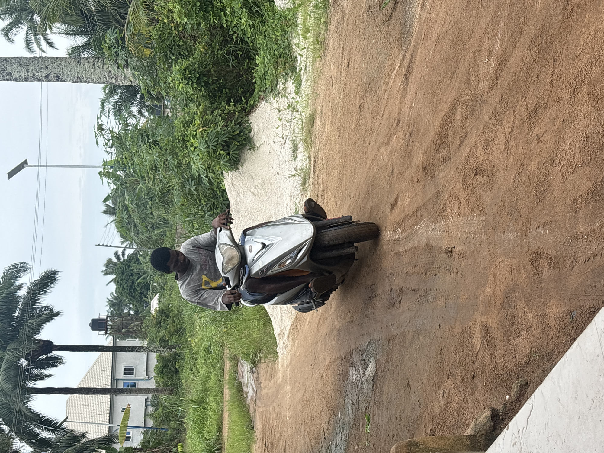 The image, set in Ikot Ekpene, Nigeria, captures a man on a scooter on an unpaved road under an overcast sky.
**Subjects:** The central subject is a man with dark skin, wearing a light-colored (possibly grey or light blue) long-sleeved shirt with some faded yellow markings on the chest, and dark trousers. He is seated on a silver and black scooter or moped, holding the handlebars. The scooter is modern in design.
**Scene and Setting:** The scene takes place on a wide, reddish-brown dirt path or road, typical of unpaved surfaces. Tire tracks are visible on the dusty ground. Lush, dense green vegetation, including numerous tall palm trees, lines both sides of the path. In the background, a light-colored house with a tiled roof is partially visible amidst the foliage. Above the house, a black water tank stands on a tall, thin structure, likely for domestic water supply. A solar-powered street light pole is visible on the left side of the path, and thin power lines crisscross the cloudy sky.
**Activity:** The man is stationary on the scooter, either having just stopped or preparing to embark on a ride. His hands are positioned on the handlebars, suggesting he is in control of the vehicle.
**Notable Details:** The sky is overcast and light grey, indicating a cloudy day without direct sunlight, likely daytime. The abundance of palm trees, tropical greenery, and the unpaved road strongly convey a tropical or subtropical environment, consistent with the specified location of Ikot Ekpene, Nigeria. There is no visible text on the man's clothing or the scooter that can be clearly read. The overall impression is of an ordinary moment in a semi-rural or residential area.