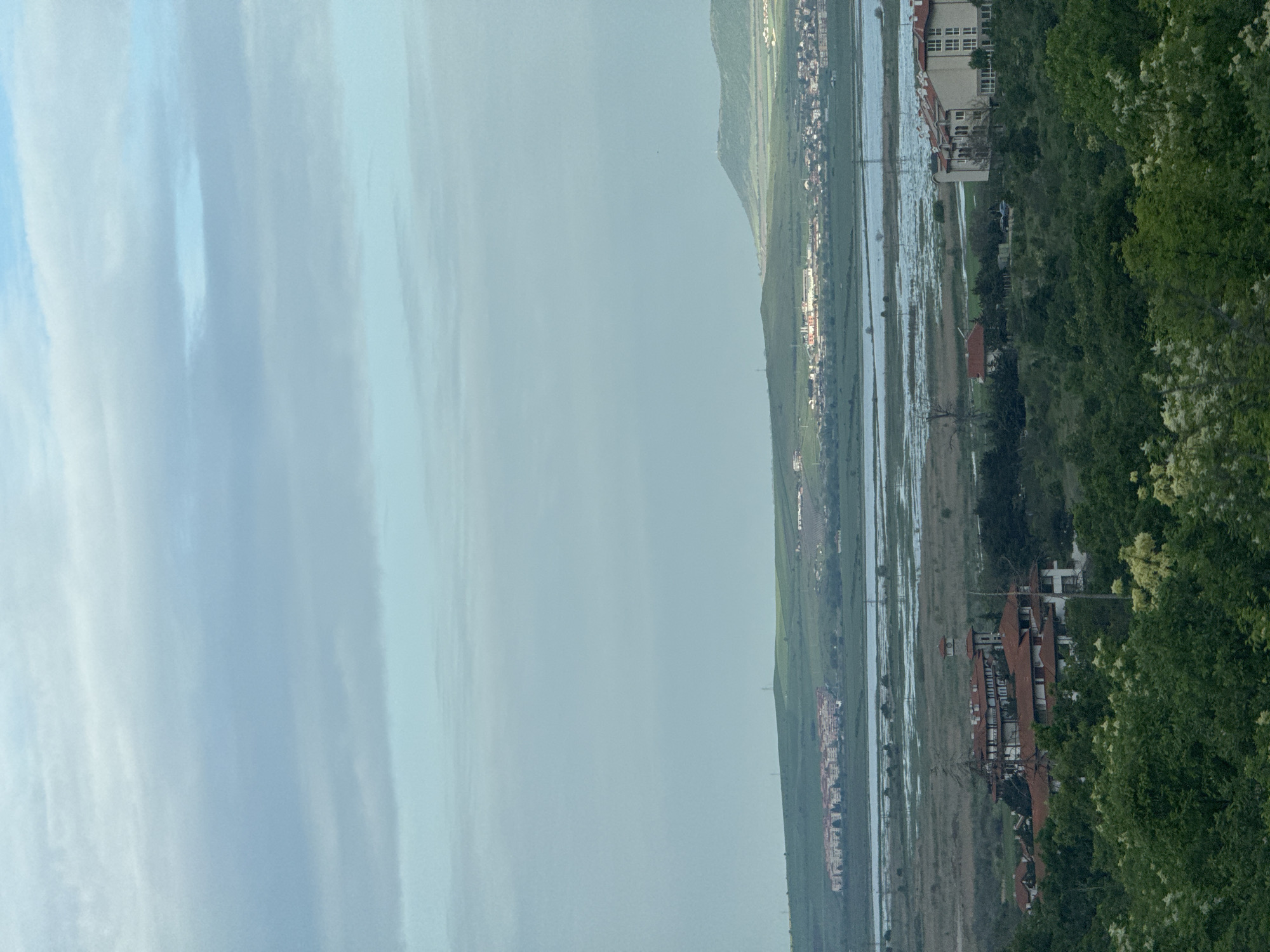 This landscape image, taken in Nesebar, Bulgaria, presents a view of the city on the Black Sea coast. To the right of the frame are trees and foliage; further from the view are buildings and houses near the coast. Behind the buildings lies the sea. Across the sea, there's a mountainous landscape with some buildings. The sky above is overcast.