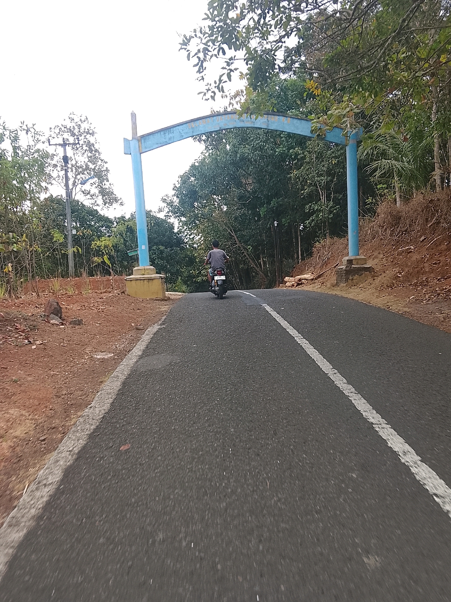 The image shows a paved road leading towards a blue archway. The archway is supported by two pillars and appears to have writing on it, though it is largely obscured by trees. A person is riding a motorcycle away from the camera, heading towards the archway. The road has a white dashed line marking the center. The surroundings are lush with green trees and vegetation, with some areas of exposed reddish-brown soil on the sides of the road. The sky is overcast. The scene appears to be a rural or semi-rural location, possibly an entrance to a park, village, or residential area. The writing on the archway suggests it is a gateway or marker. Given the context of Banjar, Indonesia, this could be an entrance to a local tourist spot or a community area.