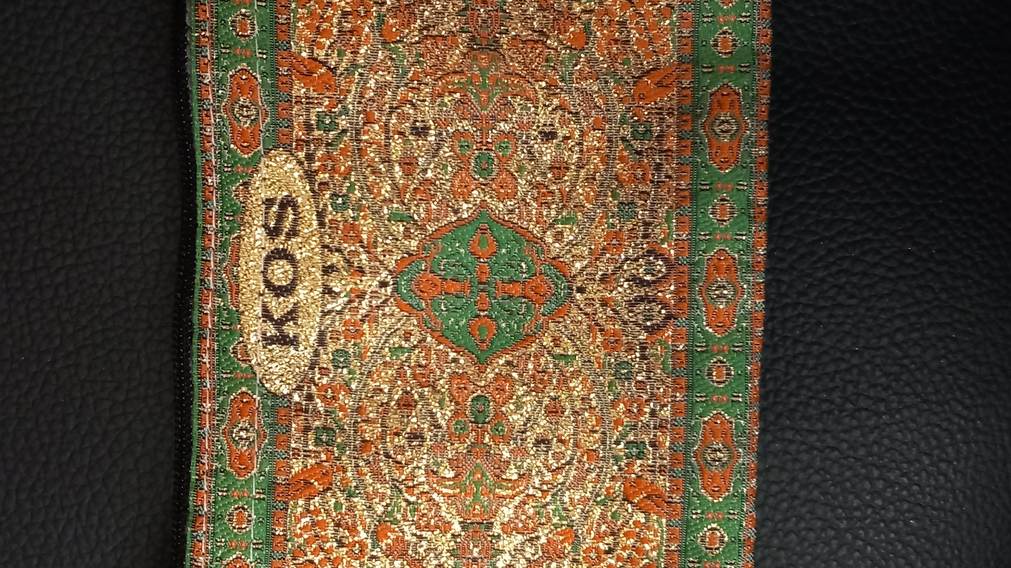 The image shows a close-up of a textured fabric with an intricate pattern. The fabric is primarily gold, orange, and green, with a prominent green border. In the center of the fabric, the word "KOS" is visible in gold, glittering letters. The fabric appears to be part of a bag or accessory, as a black zipper is visible along the top edge. The background of the image is a dark, textured material, possibly leather. There are no people or animals present in the image. The image does not provide any information about a specific location in the city of Kos, Greece, as it focuses solely on the design of the fabric.