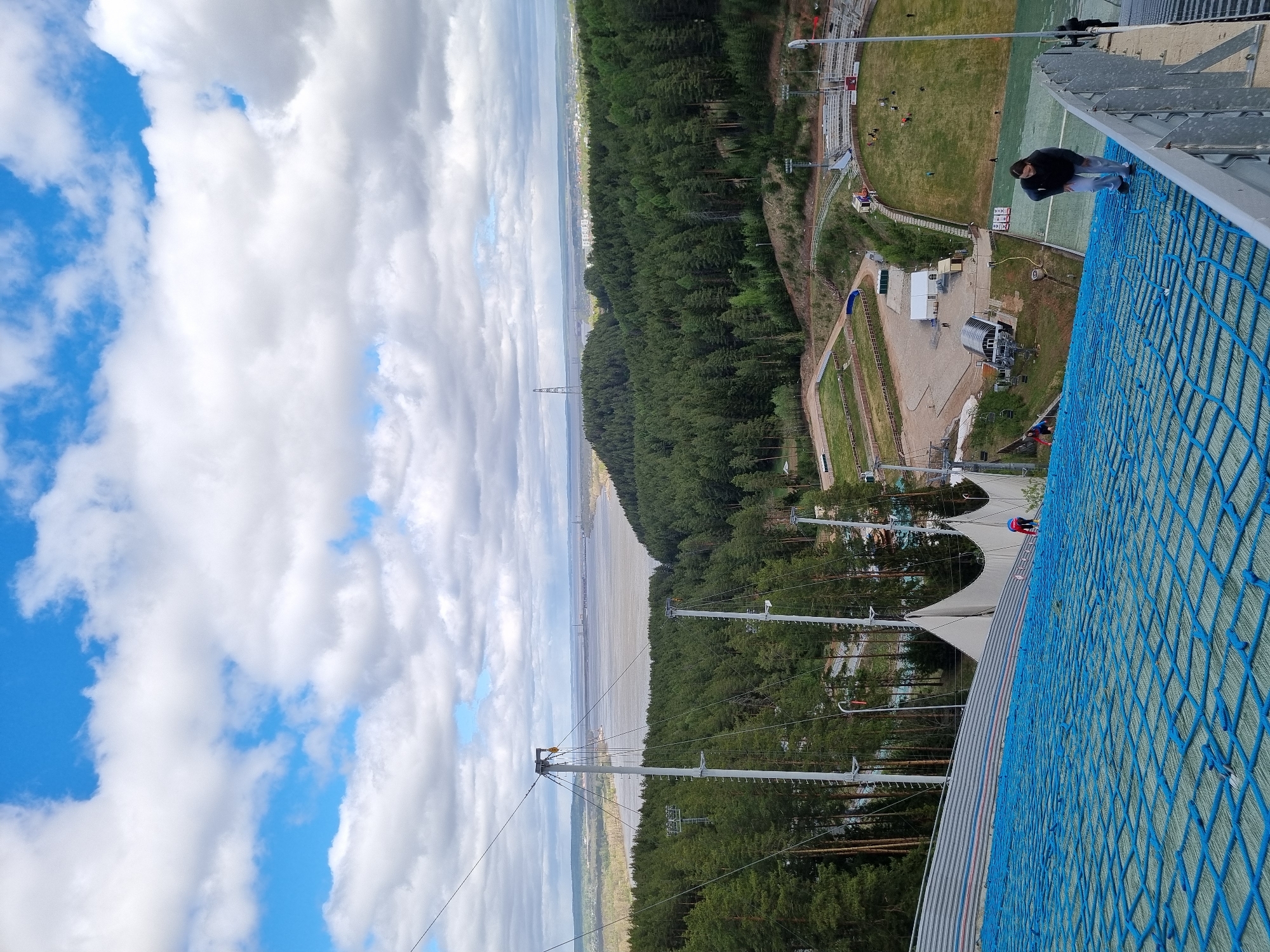 The image shows a view from the top of a ski jump in Прикамский, Russia. A man stands on the platform edge, looking down at the terrain. Below the platform, a complex of trees and other ski jump infrastructure can be seen. In the distance, there is a body of water. The sky contains a mix of blue sky and white clouds.
