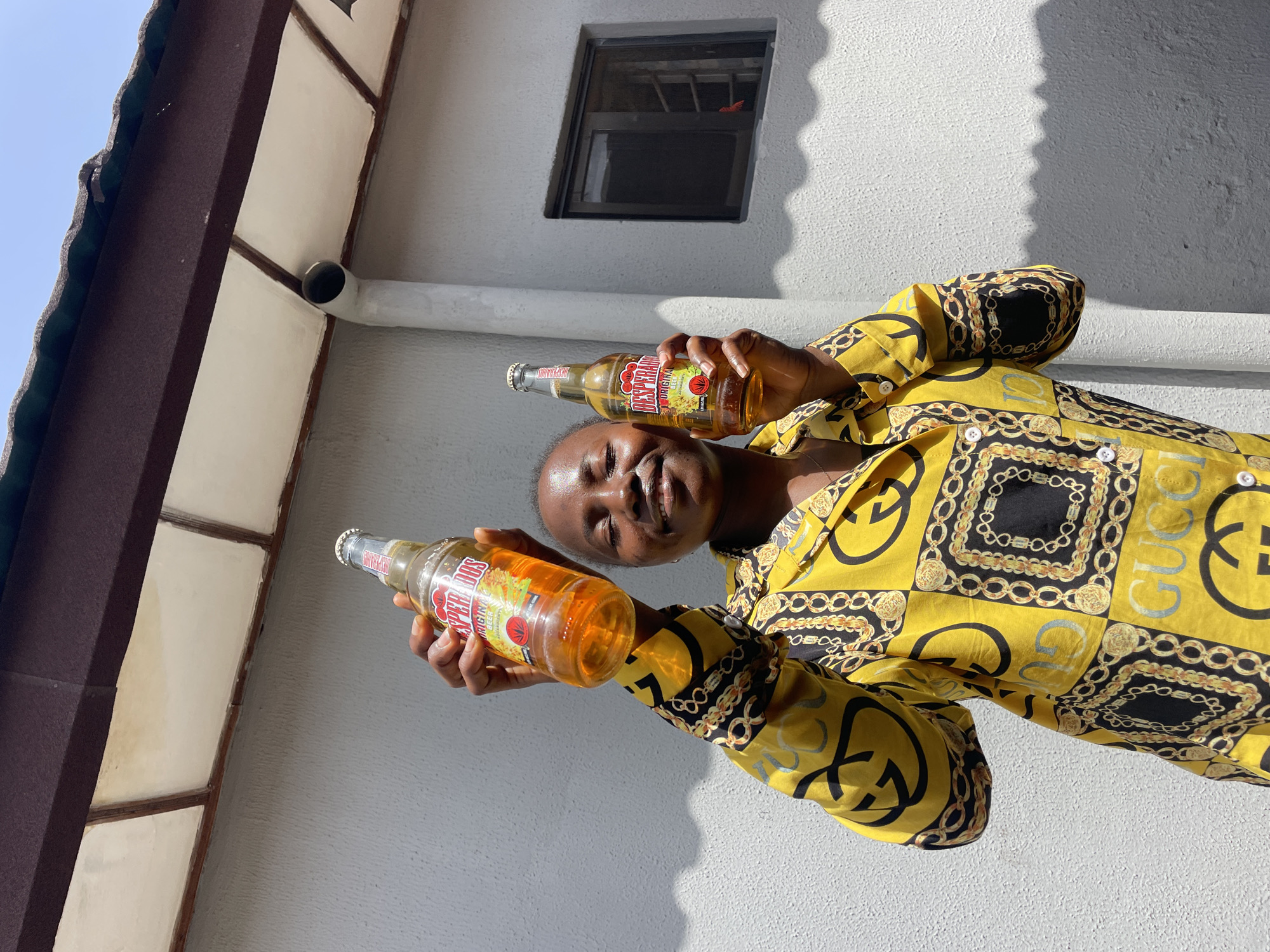 A young Black woman is holding two Desperados Original beer bottles aloft in front of a light gray stucco wall under a blue sky, with a dark brown overhang above. She is wearing a bright yellow button-down shirt with a pattern of interlocking chains, Gucci logos, and other embellishments in black and gold. The woman has her eyes closed and is smiling broadly. One bottle is in her right hand, and the other is in her left. The bottles are amber-colored, and the labels are red with white text. There is a window with dark panes in the wall to the woman's right, and a pipe runs vertically down the wall to the left of the window. The scene suggests an outdoor, casual setting, possibly a residential area, in Jos, Nigeria.