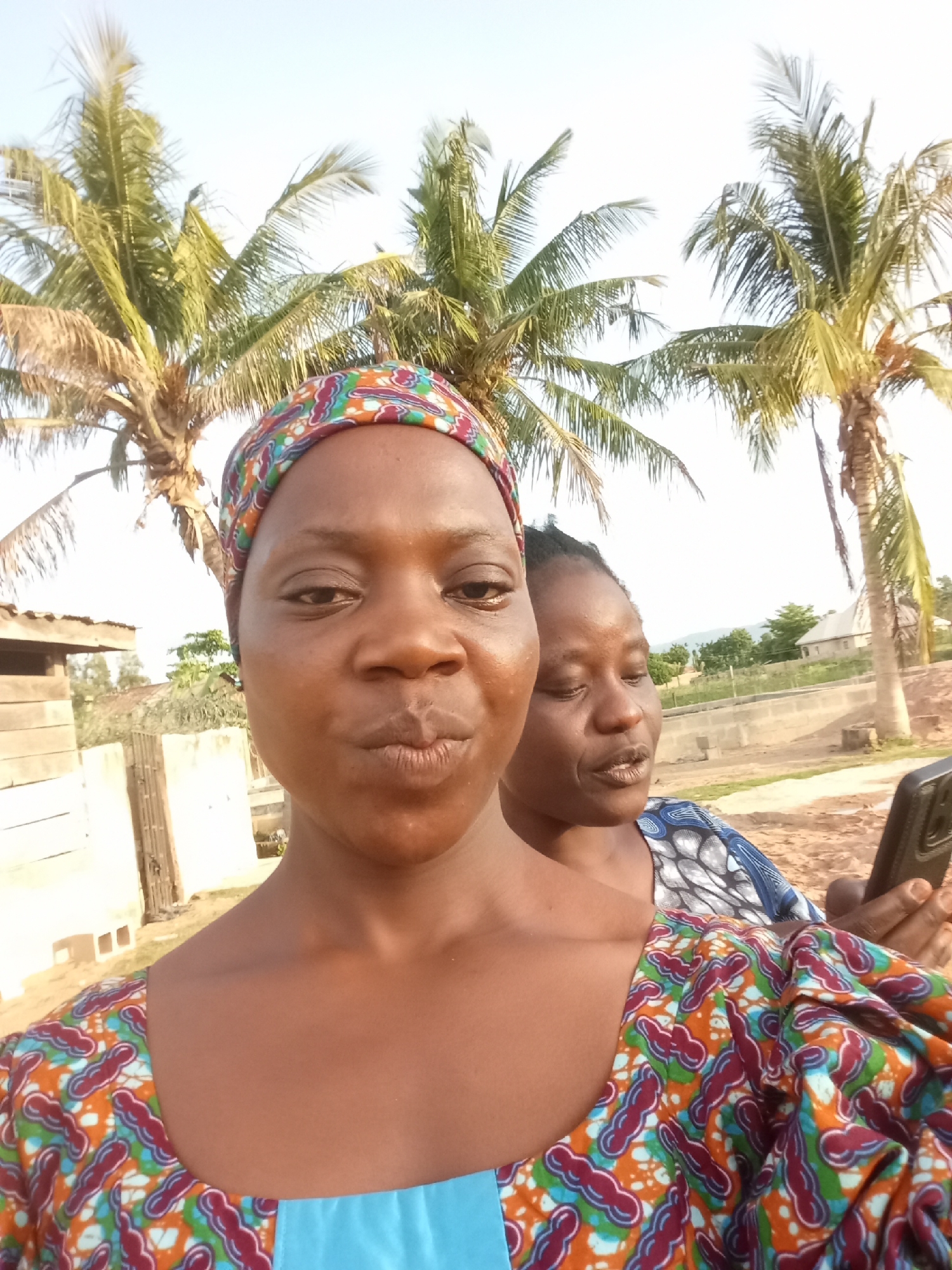 Two women are taking a selfie outdoors, with palm trees and a bright sky in the background. The woman in the foreground is looking directly at the camera, with a slight pout on her lips. She is wearing a colorful headscarf and a vibrant, patterned dress. To her right and slightly behind her, the second woman is also looking towards the camera, holding a smartphone in her right hand. She is wearing a blue and white patterned top. The setting appears to be a rural or semi-urban area, with a wooden structure visible to the left and a fence and some buildings in the background. The lighting suggests it is daytime, possibly late afternoon given the warmth of the sunlight. The overall mood seems relaxed and informal, consistent with a casual selfie.