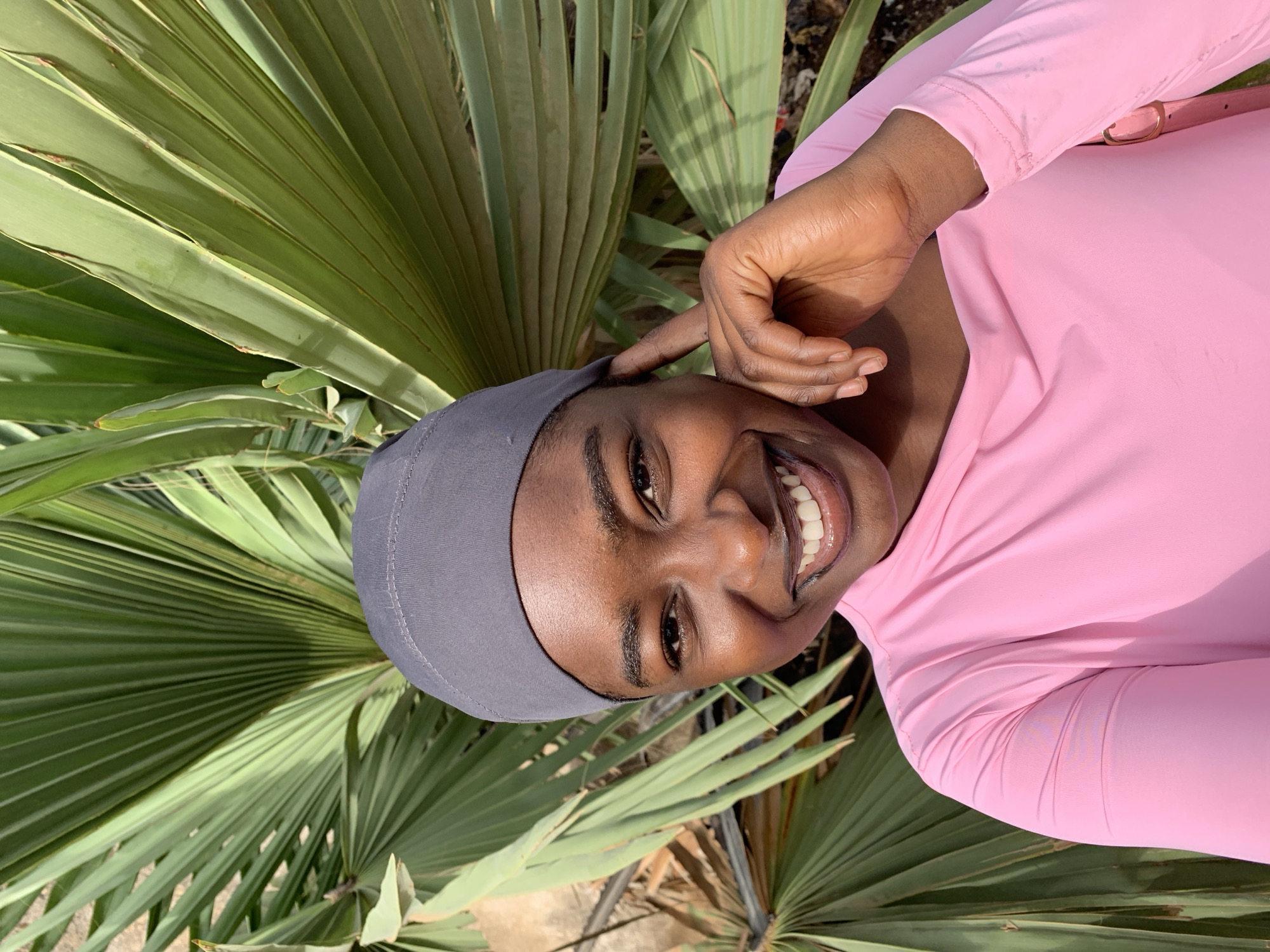 The image shows a woman with dark skin and a wide smile, wearing a gray head covering and a pink long-sleeved shirt. Her hand is near her head. She is positioned in front of a large green palm-like plant with fan-shaped leaves. The location is outdoors, presumably in the city of Jos, Nigeria, as requested. The background details are minimal.