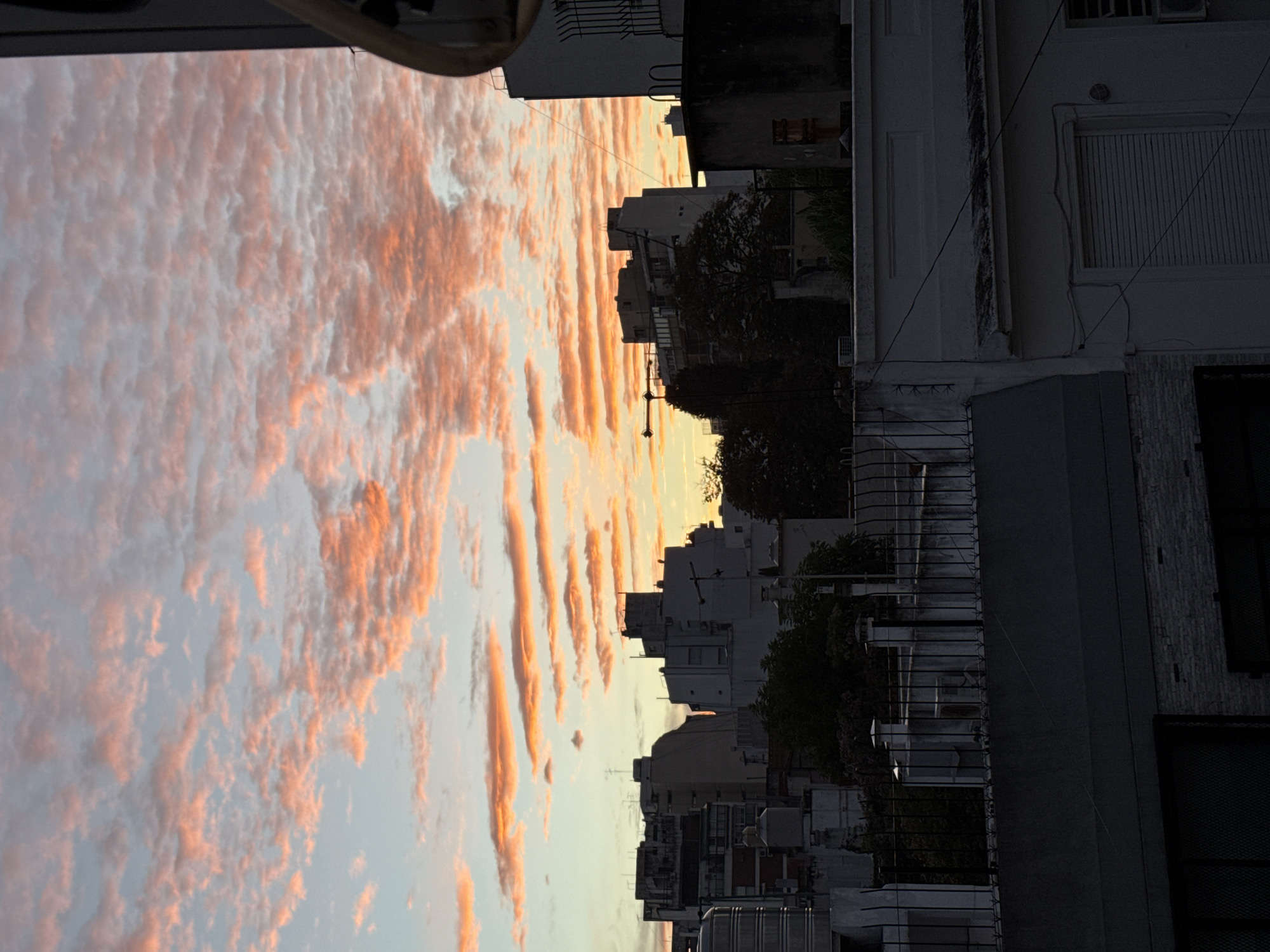 The image captures a sunset scene in Buenos Aires, Argentina, from a street-level perspective looking upwards. The foreground features buildings with various architectural styles, including balconies with metal railings, windows with shutters, and visible electrical wiring. In the background, there is a clear separation to the sky with rows of orange-hued altocumulus clouds, which dominates a significant portion of the frame. The overall tone of the image is calm and urban, with the soft light of the sunset contrasting with the darker shades of the building exteriors. No people or visible human activity are discernible in the frame.