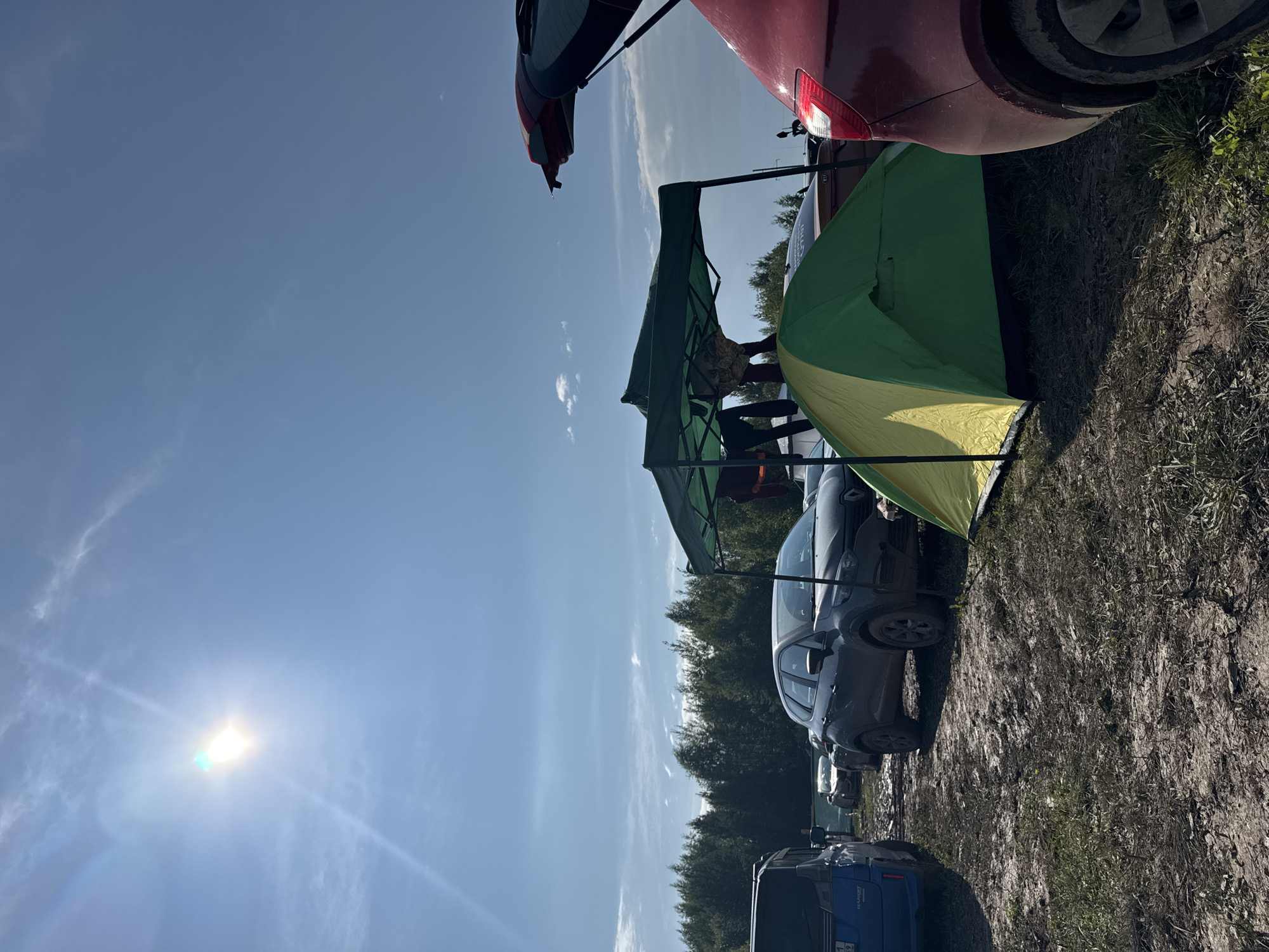 The image depicts an outdoor scene, likely a campsite, with several vehicles and camping equipment. The ground is muddy and appears to be dirt with some patches of grass. In the foreground, a green and yellow tent is pitched, and a portion of a red car with its trunk open is visible on the right. Behind the tent, a dark grey car is parked. A green canopy tent is set up over some items, and a person is partially visible standing under it. To the left, a blue van is parked. In the background, a dense line of evergreen trees forms a treeline against a bright blue sky with scattered clouds. The sun is shining brightly in the upper left corner of the image, creating lens flare. The scene suggests a temporary outdoor gathering or event. The presence of multiple vehicles and camping gear implies a recreational or temporary living situation. The environment is rural or semi-rural, given the treeline and the unpaved, muddy terrain.