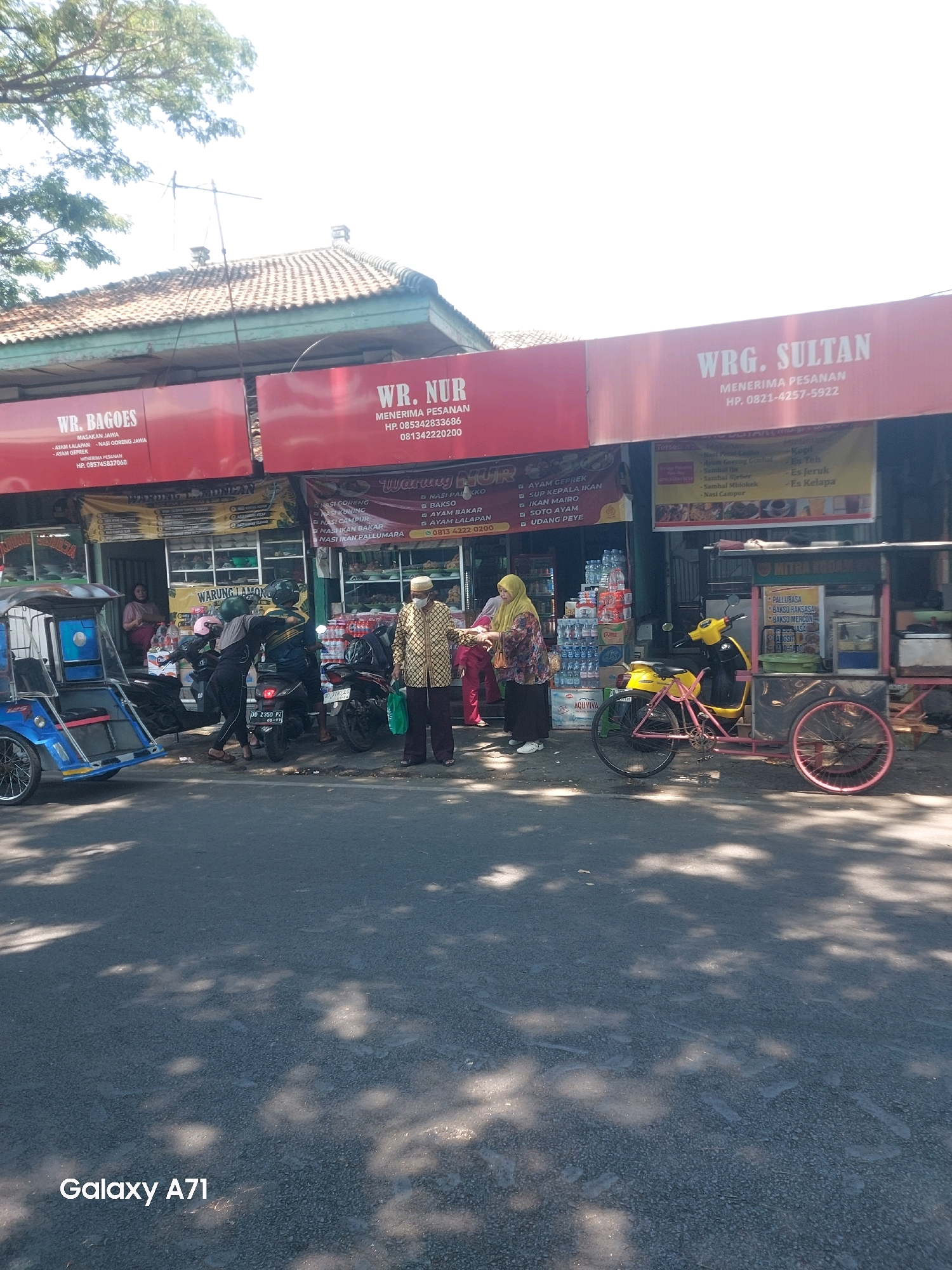 The image shows a street scene in Makassar, Indonesia, with several storefronts and food stalls. On the left, "WR. BAGOES MASAKAN JAWA" is advertised. To its right, "WR. NUR MENERIMA PESANAN" is displayed, with phone numbers. Further to the right, "WRG. SULTAN MENERIMA PESANAN" also shows contact information. In front of the stores, people are interacting, with some standing and others on motorcycles. A blue and white pedicab is parked on the left. A yellow and pink food cart with a motorcycle is on the right. Bottles of water and other goods are displayed for sale. The street is paved, with shadows from trees falling on the surface. A watermark at the bottom reads "Galaxy A71".