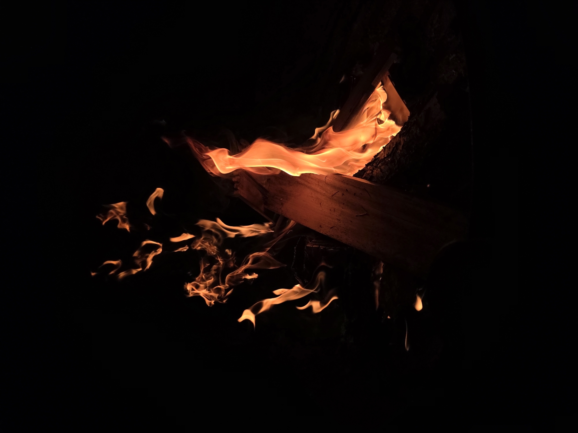 A campfire burns in the dark. The flames are a vibrant orange and yellow, licking at the dark wood. The scene appears to be in the city of Строитель-нефтяник Снт, Russia. The fire is the central focus of the image, with the dark background suggesting it is nighttime or a dimly lit area. There are no people or animals visible in the image.