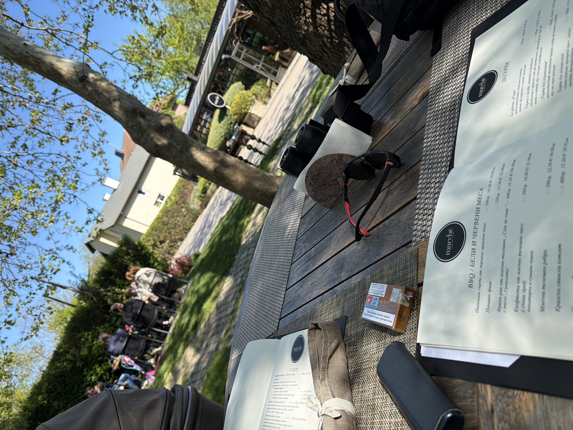 The image depicts a partially occupied outdoor dining table in what appears to be a restaurant setting. On the table, there are menus from "marché", cigarettes, sunglasses, a small camera, a coaster, a napkin, and a backpack. In the background, one can see a restaurant structure, trees, a building, and people sitting. The menus indicate the restaurant is offering BBQ, white and red meats, and desserts. The text on the menu is in Bulgarian. The restaurant appears to be located outside of a park and is accessible from a side street. The location is likely Varna, Bulgaria, based on the language used.
