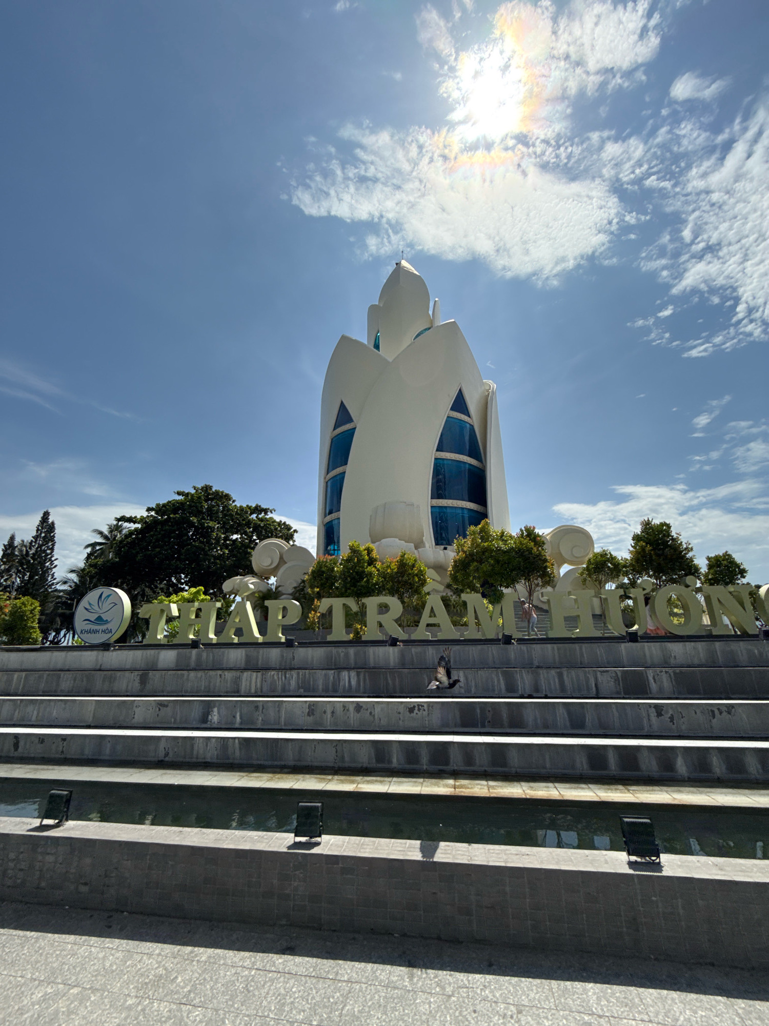 The image shows the Po Nagar Cham Towers in Nha Trang, Vietnam. The towers are made of red brick and are located on a hill overlooking the city and the sea. The sky is blue with some white clouds. In the foreground, there are some steps leading up to the towers. There are also some trees and bushes. The overall scene is one of peace and tranquility. The text in the image reads "THAP CHAM HUONG" which is the Vietnamese name for the towers.