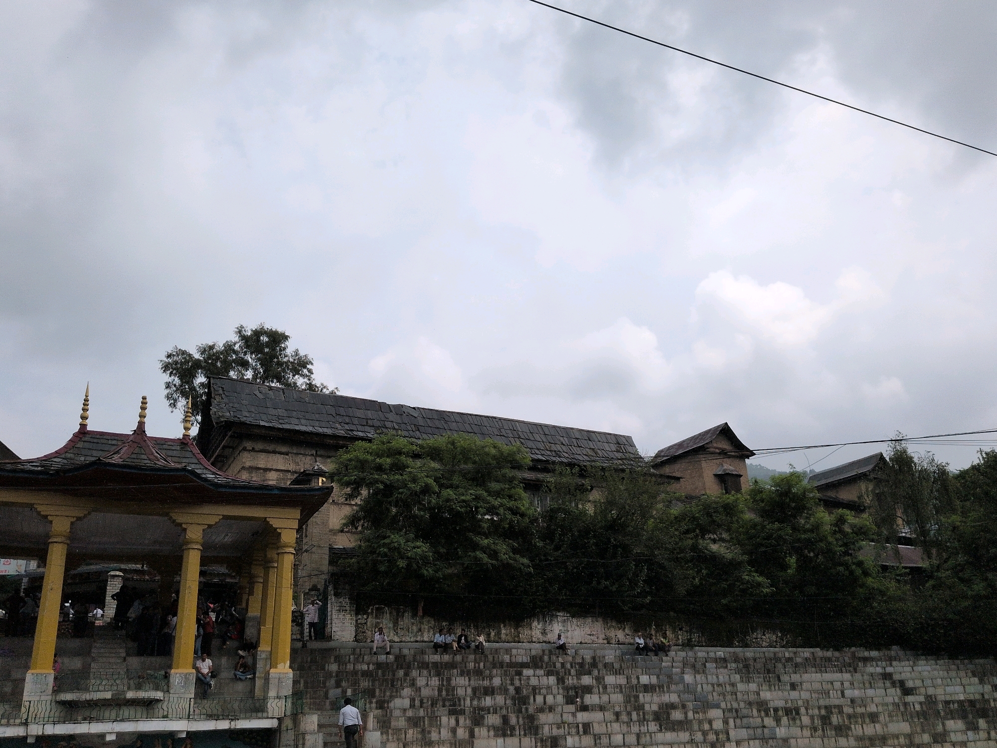 The image depicts the ghats along the Beas River in Mandi, India. On the left, a colorful pavilion with yellow pillars and a dark, tiered roof stands prominently, with people gathered underneath and on the steps. Behind the pavilion, a series of stone buildings with dark, sloping roofs are visible, partially obscured by dense green foliage. The buildings appear to be made of stone or brick and have a rustic, traditional architectural style. A grey stone retaining wall with a textured surface stretches across the foreground, with several people sitting on its steps. The sky is overcast with a soft grey hue, suggesting cloudy weather. A thin black wire runs diagonally across the upper right portion of the frame. The overall scene conveys a sense of everyday life and local architecture in Mandi.
