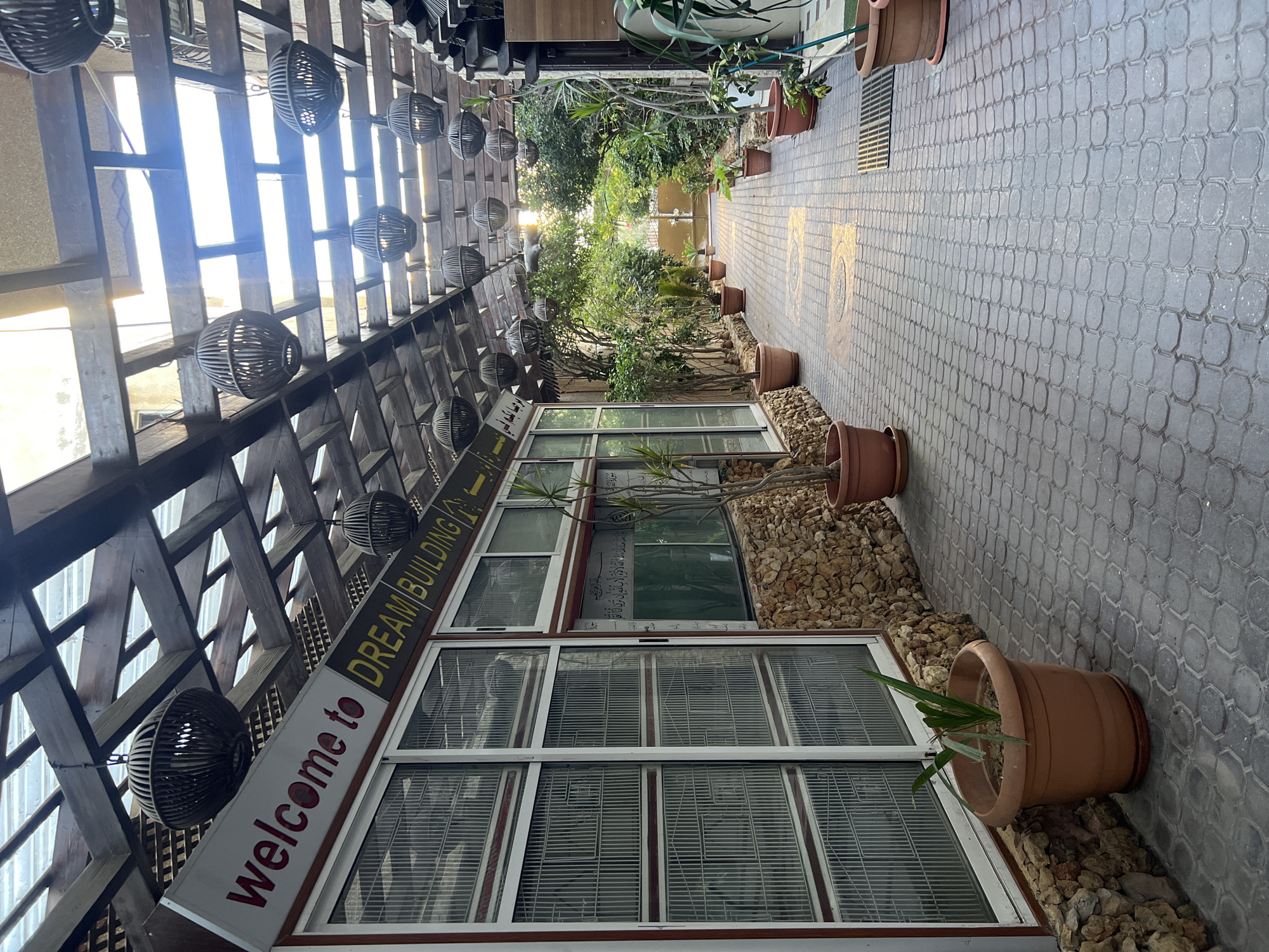 The image depicts a covered walkway in Gaza City, Palestinian Territories, with a traditional Arabic architectural style. The walkway is paved with hexagonal interlocking stones, and the roof is constructed of wooden beams and translucent panels, allowing sunlight to filter through. Hanging from the wooden beams are spherical decorative lanterns made of woven material.
On the left side of the walkway, there is a building with large windows featuring metal grates. A sign above the windows reads "welcome to DREAM BUILDING" in red and yellow lettering. The building's facade is decorated with rough-hewn stones. Potted plants are placed along the walkway.
To the right of the walkway, lush green trees and plants create a natural barrier, leading towards a gate in the distance. The overall scene suggests an outdoor public space, possibly an entrance to a commercial or residential complex, characterized by its blend of natural elements and built structures. The lighting indicates it is daytime, possibly late afternoon due to the warm glow of the sun.
