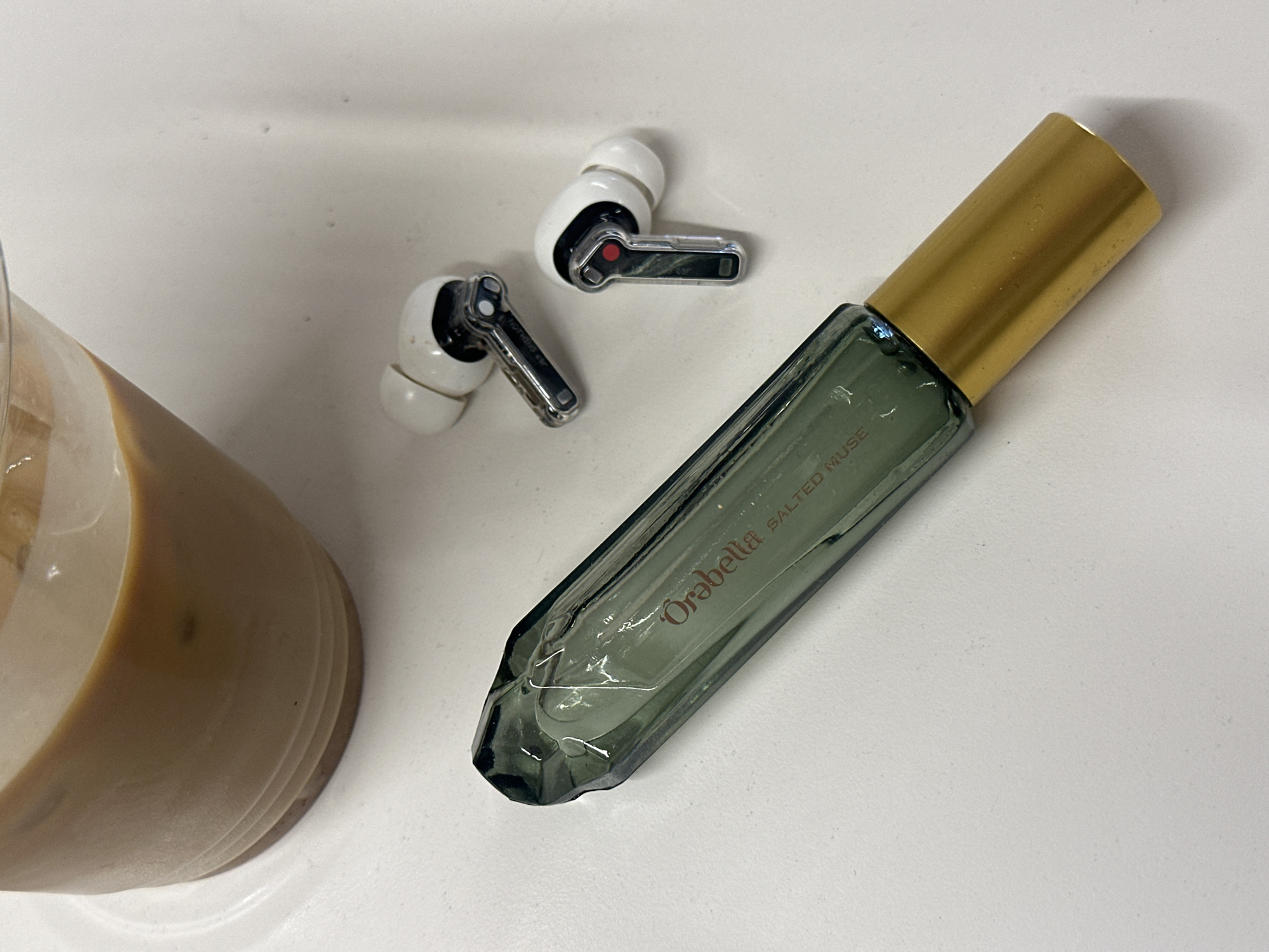 The image shows a white surface with a glass of liquid, a pair of white earbuds, and a green perfume bottle with a gold lid. The perfume bottle has the text "Orabella SALTED MUSE" printed on it. The glass is partially filled with a beige liquid, possibly coffee or a latte. The earbuds are white with silver detailing. The scene does not show a location in London, United Kingdom.