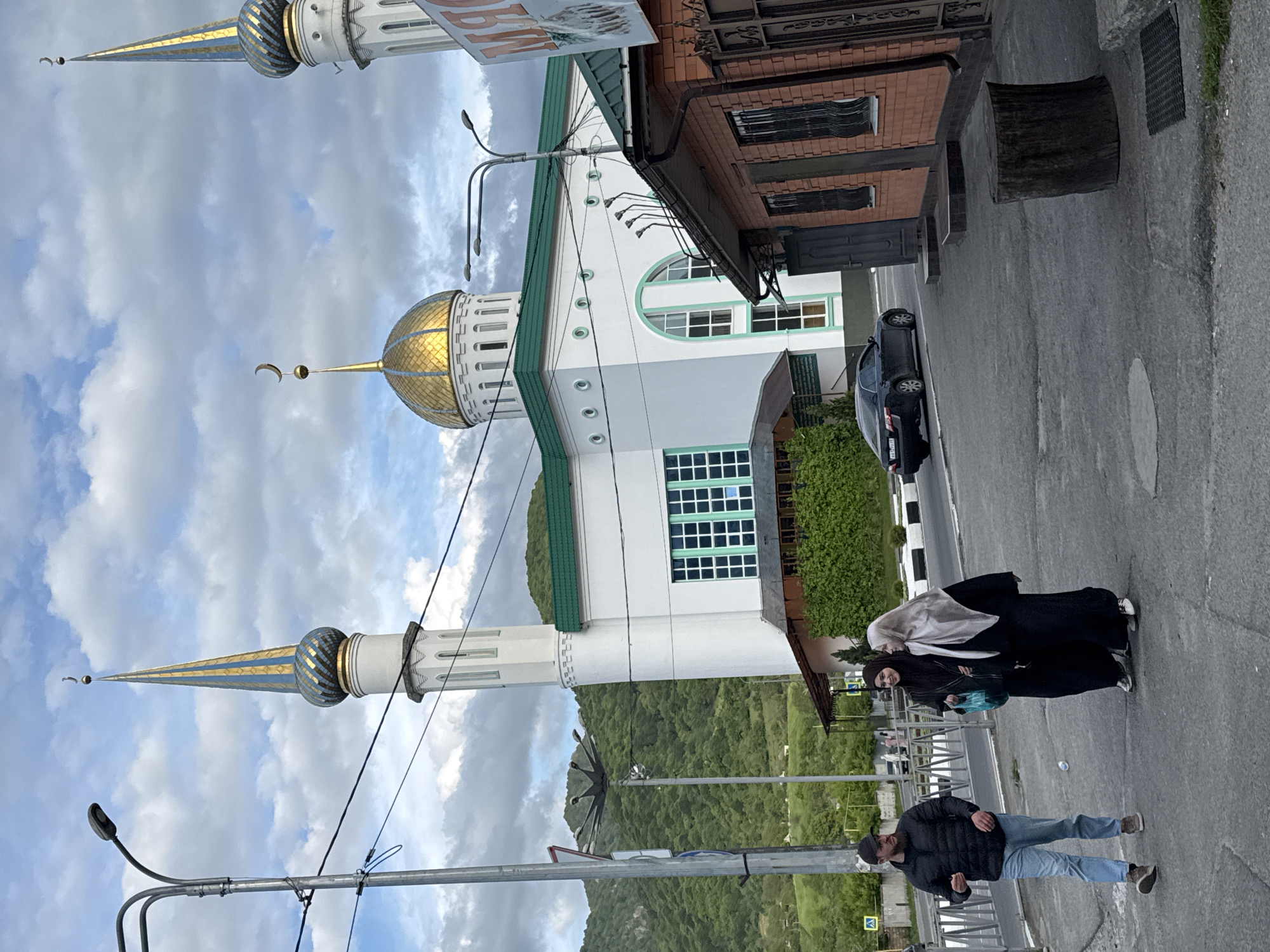 The image, which is presented rotated, depicts a vibrant street scene in Karachayevsk, Russia, featuring a mosque and forested mountains under an overcast sky.
In the midground, a prominent white mosque with a green roof stands out. It features a large golden dome adorned with a crescent moon finial and two tall, slender minarets, each capped with a golden and blue patterned spire and a crescent moon. The mosque's facade includes both arched and rectangular windows. Adjacent to the mosque on its right is a multi-story brick building with dark windows.
An asphalt road occupies the foreground and midground. A dark-colored car is parked on the side of the road near a curb marked with black and white stripes. On the left side of the road, a man wearing a dark jacket, blue jeans, and a baseball cap is walking towards the viewer. Further to the right, two women are standing; one is dressed in a light-colored hijab and a black abaya, while the other wears dark headwear and clothing.
Lush, green, forested mountains form the backdrop, rising under an overcast sky with patches of white clouds and blue visible. Streetlights, utility poles, and wires are present along the road, indicating urban infrastructure. The golden dome and intricate spires of the minarets are notable architectural details. There is no clearly legible text visible on any signs.