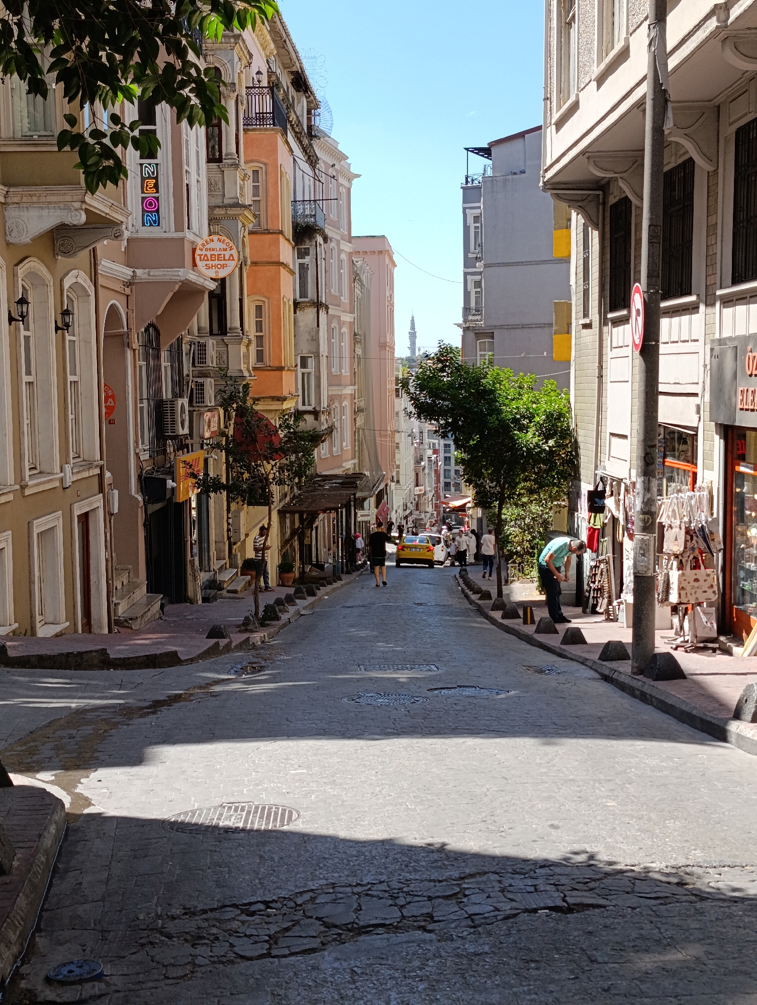 The image depicts a narrow, cobblestone street in Beyoğlu, Turkey, lined with multi-story buildings featuring colorful facades and intricate architectural details. The street slopes gently downhill, with a yellow taxi visible in the distance. Several people are walking on the sidewalks, engaged in various activities, such as shopping and browsing. One man is bent over, examining items displayed outside a shop. The scene is bathed in bright sunlight, casting distinct shadows on the street and buildings. The architecture suggests a historical urban setting, characteristic of neighborhoods like Beyoğlu, known for its charming streets and vibrant atmosphere.