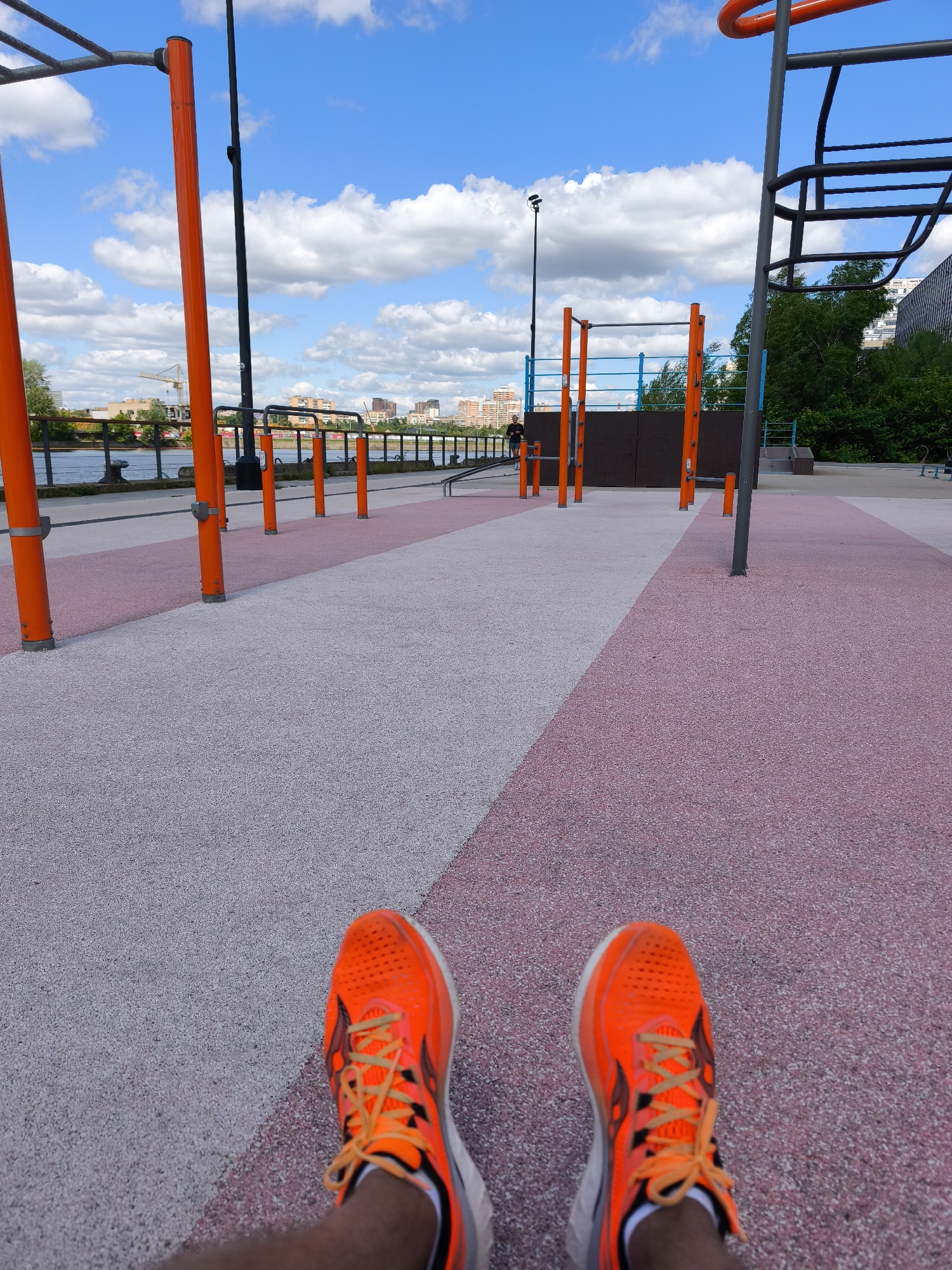 A first-person perspective shot shows bright orange running shoes on a person's feet. The shoes are laced and appear to be athletic footwear. They are positioned on a ground surface that is a mix of light grey and pink speckled material, possibly a rubberized or composite surface often found in outdoor recreational areas.
In the background, an outdoor fitness park is visible with several orange poles and metal bars designed for exercise. A person can be seen in the distance near some of the equipment. Beyond the park, a body of water, likely a river, is present, with a waterfront promenade and railing. In the far distance, a cityscape with buildings, a crane, and trees can be observed under a bright blue sky with scattered white clouds. The scene suggests a sunny day at an urban park. The location is identified as an outdoor workout area near the Moscow River embankment, Russia.