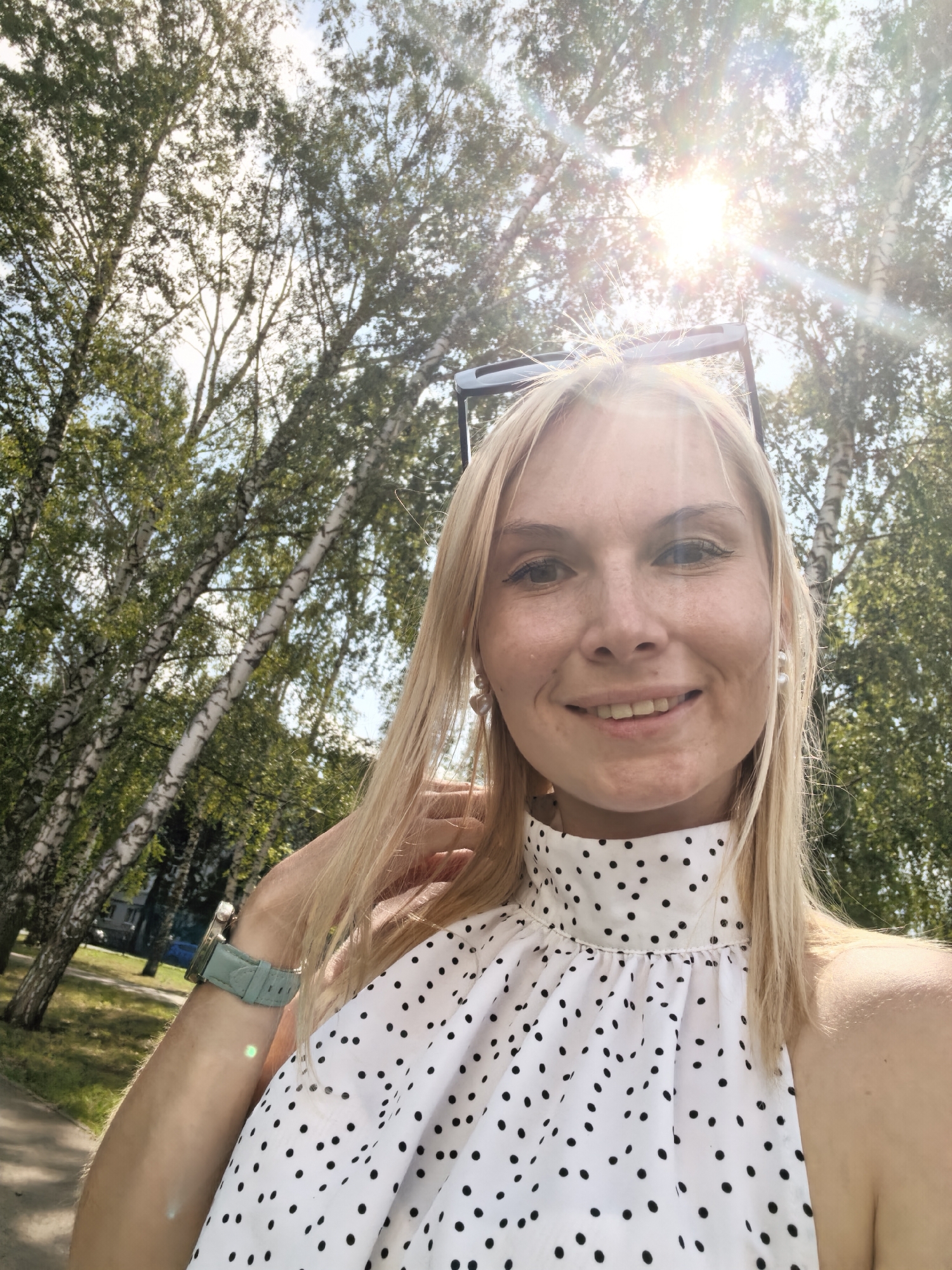 A blonde woman is taking a selfie outdoors with a bright sun shining through the trees. She is smiling and wearing a white top with black polka dots, pearl earrings, and a light blue watch on her left wrist. The background shows a park with birch trees and a path. The woman is outdoors in the city of Sterlitamak, Russia.