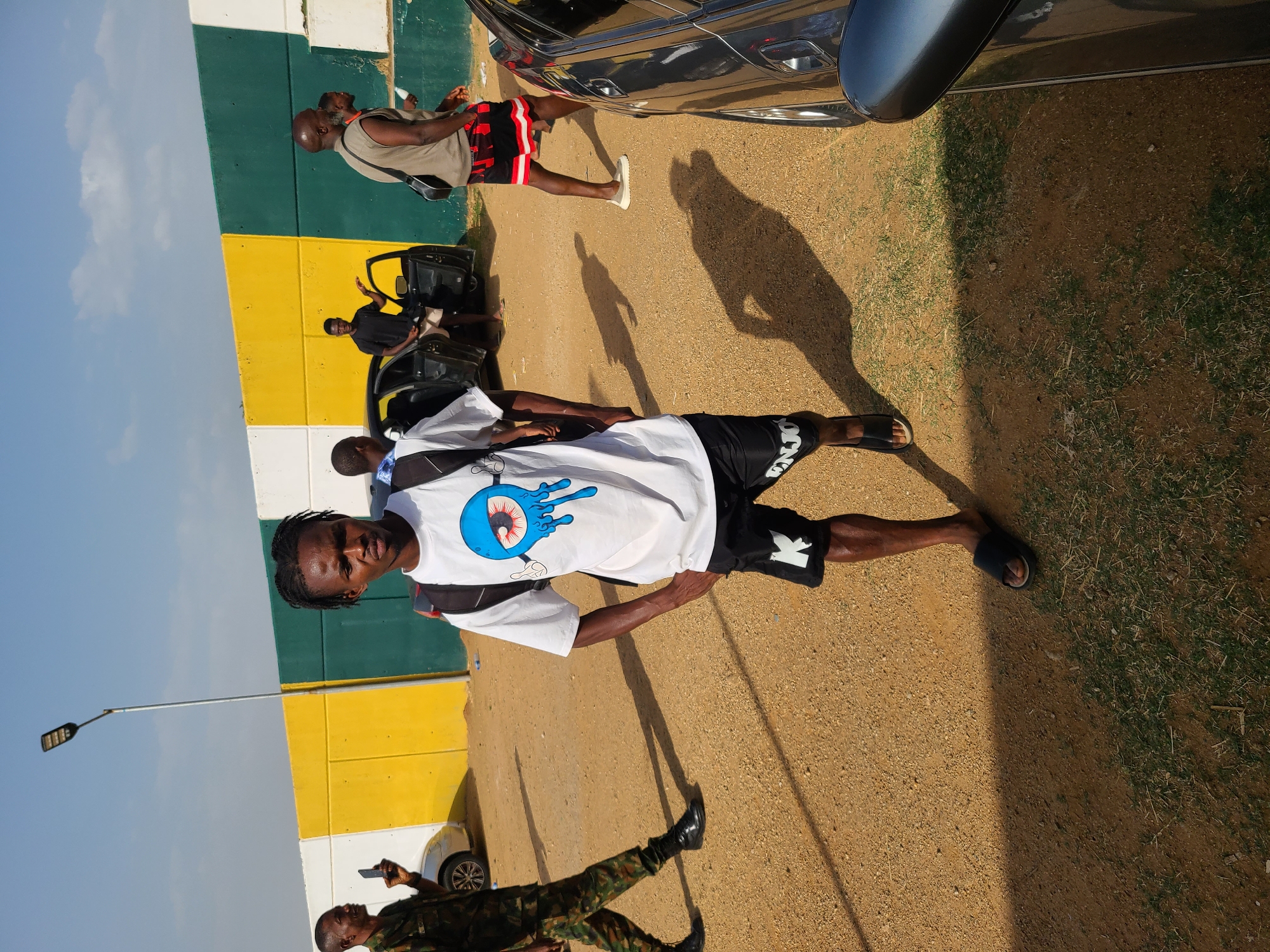 The image depicts an outdoor scene, possibly in or near New Stadium, Nigeria, given the presence of individuals and details suggesting the location.
**People:**
- A young man is standing in the foreground, wearing a white t-shirt with a blue graphic design, black shorts, and sandals. He has a backpack on his back.
- Another man, dressed in military camouflage uniform, is walking in the lower-left of the frame.
- Other individuals are visible in the background, some near parked vehicles. One man is wearing a beige tank top and red and black patterned shorts. Some carry bags.
**Objects:**
- Several vehicles are present, including a dark-colored car in the upper part of the frame.
- Bags, potentially luggage, are located in the background.
- A street light is on the left side of the image.
**Actions/Interactions:**
- The main subject is posing for the picture.
- People in the background are engaged in various activities, suggesting movement or transportation.
**Environment:**
- The ground is a dirt or gravel surface.
- There is a wall painted in horizontal stripes of white, yellow, and green in the background.
- The sky is visible in the upper left corner and is partially clear.