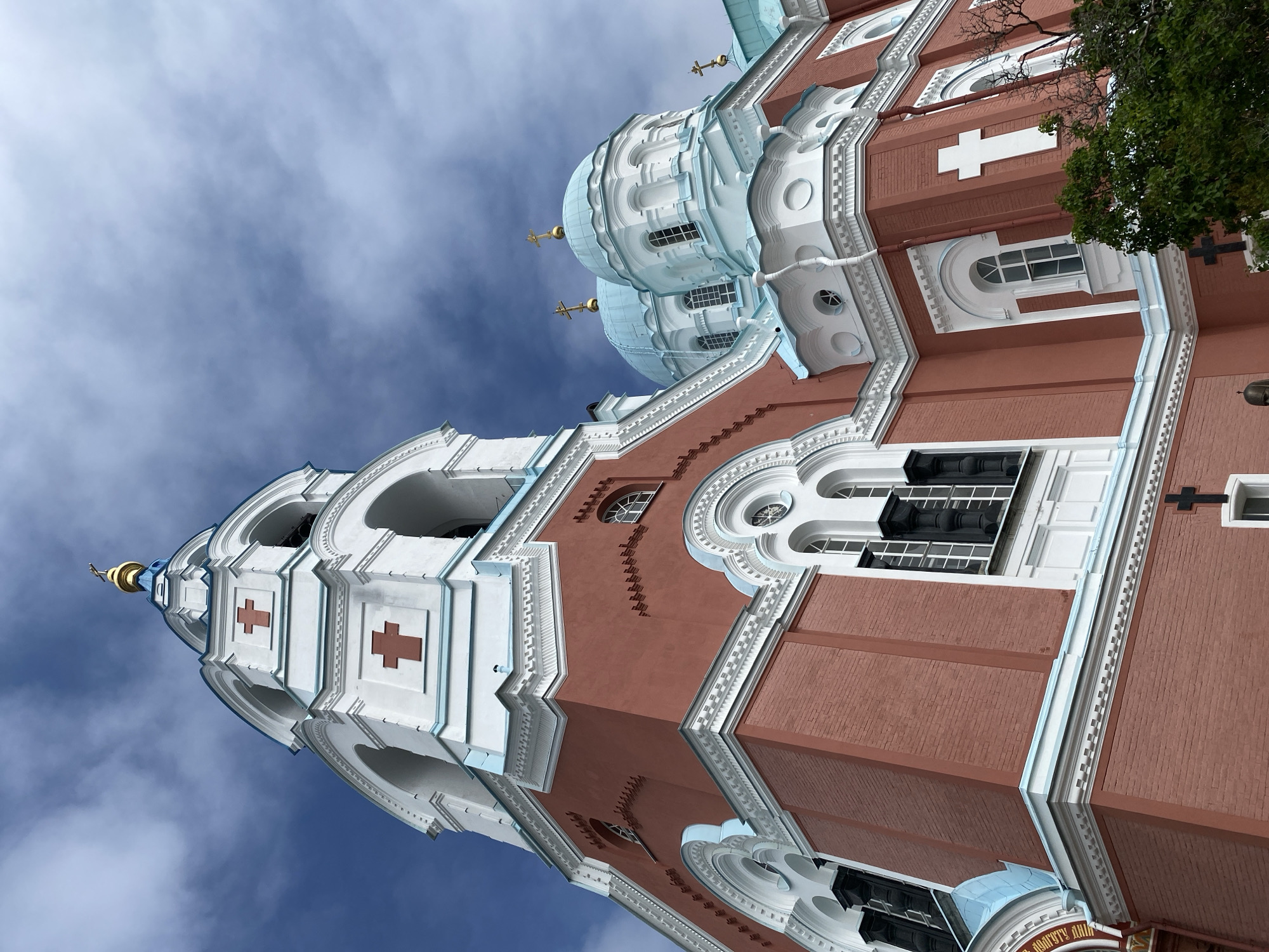 The image shows a low-angle, full-frame shot of the Transfiguration Cathedral on Valaam Island, Russia. The cathedral is a grand, red-brick building with white architectural details and light blue domes topped with golden crosses. A prominent bell tower with arches and red crosses on its white facade is visible on the left. The sky above is partly cloudy with blue sky peeking through. The architecture appears to be of Russian Orthodox design. The image is a static exterior shot with no people or animals present.
