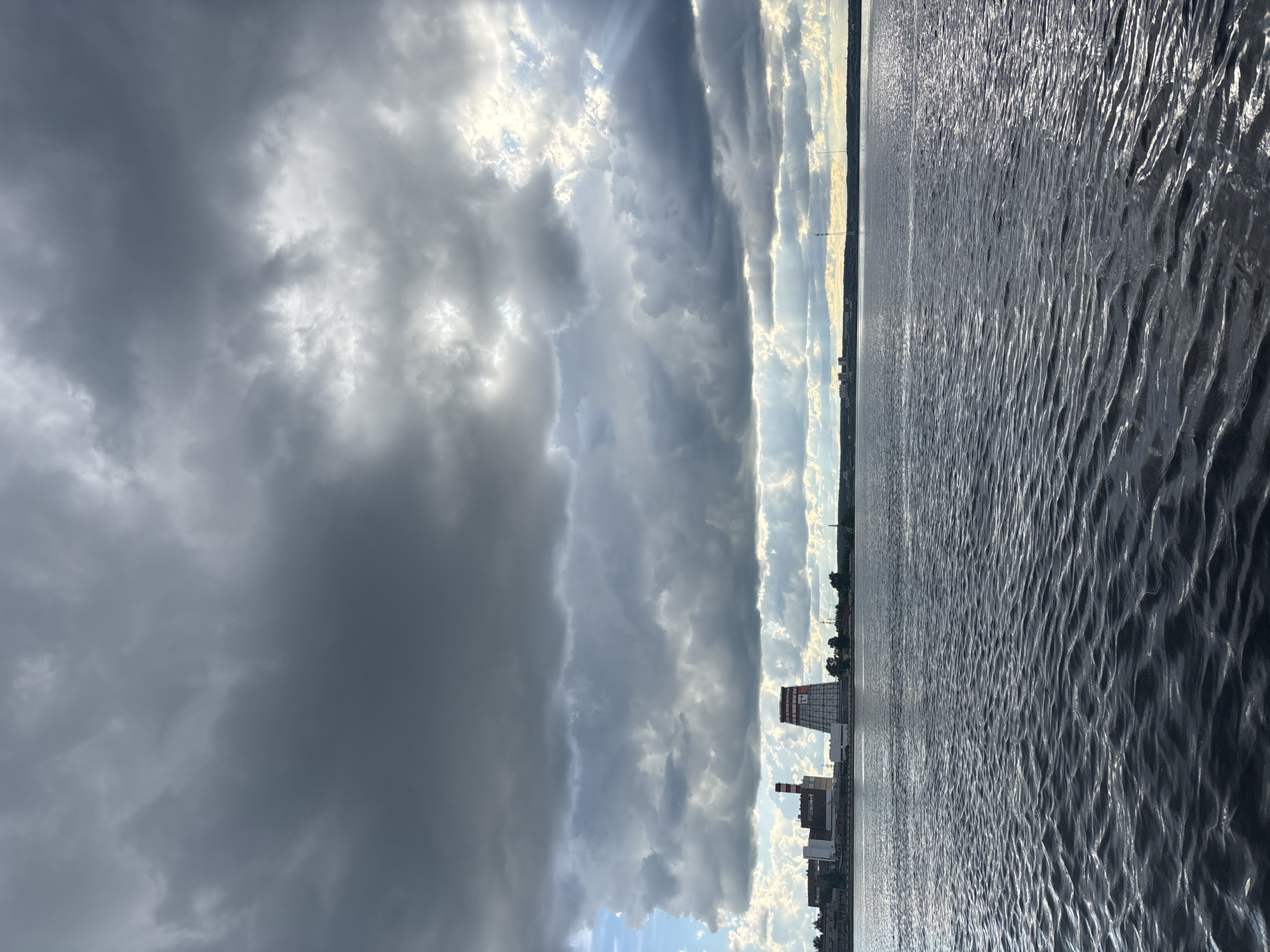 The image captures a landscape with a dramatic sky and water. On the left, a dark, heavy cloud formation dominates the sky, contrasting with the brighter, sun-dappled clouds towards the center. Vertical shafts of light break through the cloud cover. To the right, the water is choppy with light reflecting off the surface. Along the horizon, a city skyline is visible, including industrial buildings, specifically what appears to be a power plant identified as Izhevsk CHPP-2, in the Russian city of Izhevsk. The image is oriented vertically, presenting the landscape in an unusual aspect ratio.