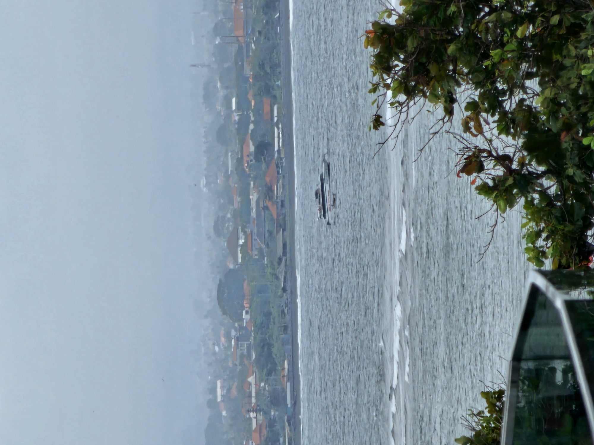The image depicts a hazy coastal scene, likely in Denpasar, Indonesia, characterized by a dense, low-lying urban sprawl extending to the horizon under a muted sky. The foreground features a railing and lush green foliage, suggesting a vantage point from an elevated structure overlooking the sea. The ocean itself is a turbulent expanse of gray-blue water, with visible whitecaps indicating choppy conditions. Several boats are present on the water; a larger vessel, appearing to be a catamaran or ferry, is positioned towards the center, with smaller boats scattered nearby. The town beyond the shore is densely packed with buildings, many featuring characteristic terracotta-colored roofs, interspersed with trees. The overall atmosphere is one of a humid, overcast day.