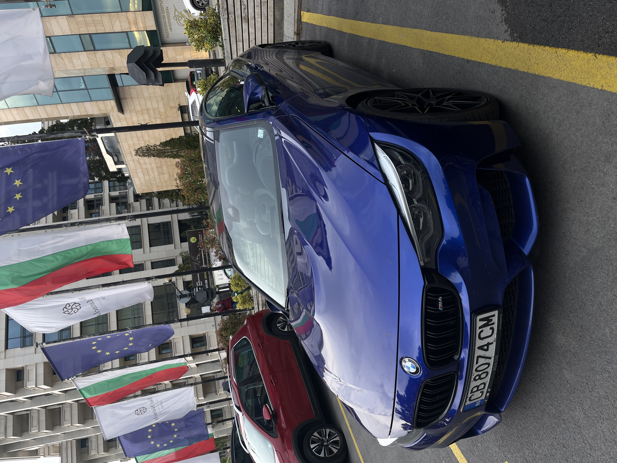 The image, rotated vertically, depicts an urban street scene in Sofia, Bulgaria, featuring parked cars and a display of flags. In the foreground, a vibrant blue BMW M series car is prominently parked on asphalt, outlined by a yellow painted line. The car's front grille, adorned with the BMW logo and an 'M' badge, and its sleek headlights are clearly visible. Its Bulgarian license plate reads "BG CB 8074 CM", with "CB" indicating Sofia registration, and also includes the text "NOVA CAR".
Behind the blue BMW, a red car, likely an SUV or crossover, is partially visible. The background is dominated by modern, multi-story buildings with light-colored stone facades and numerous reflective glass windows. Along the street, a series of flags are displayed vertically on poles. These include the national flag of Bulgaria (white, green, and red horizontal stripes) and the flag of the European Union (blue with a circle of yellow stars). Interspersed among these are white flags, each featuring a distinct black, stylized knot-like logo and Cyrillic text. On one white flag, the text "ЕЛЕОНОРА" is legible. On a building facade, Cyrillic text "МЕДИЦИНСКИ ЦЕНТЪР" (Medical Center) and a possible name like "ФЛОРИНЕЛ" can be seen.
A black, abstract, zigzag-shaped sculpture or sign is erected on a pole near the buildings. The scene takes place during the daytime under bright, possibly overcast conditions, evidenced by the reflections and general illumination. A few indistinct human figures are visible in the background, near the buildings and the red car, suggesting general urban activity.