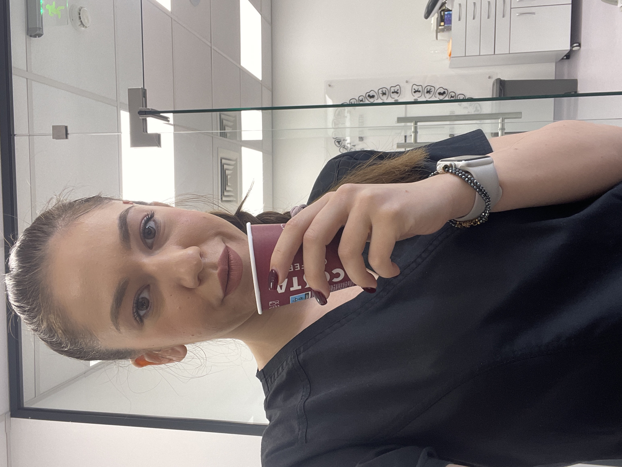 A young woman is captured in a well-lit indoor setting, likely a modern dental or medical office. She appears to be in her 20s or early 30s, with light skin, dark hair pulled back, and dark eyes. She wears a black scrub-style top. On her left wrist, she has a white smartwatch with a grey band, accompanied by a beaded bracelet. Her right hand holds a disposable Costa Coffee cup, with her fingernails painted a dark, possibly maroon, color. She looks directly at the camera with a subtle, confident smirk.
The background reveals a clean, contemporary environment. Clear glass partitions divide spaces, reflecting bright rectangular overhead lights. On a white wall behind her, medical cabinetry is visible. Most notably, a clear acrylic plaque displays diagrams of teeth, clearly labeled "UPPER TEETH" and "LOWER TEETH," strongly indicating a dental practice. A small white sensor and a green light with a figure (possibly an exit sign) are also mounted on the wall. The text "COSTA COFFEE" is visible on the cup she holds. The overall impression is a professional but relaxed moment during a workday in a dental office located in Sofia, Bulgaria.
