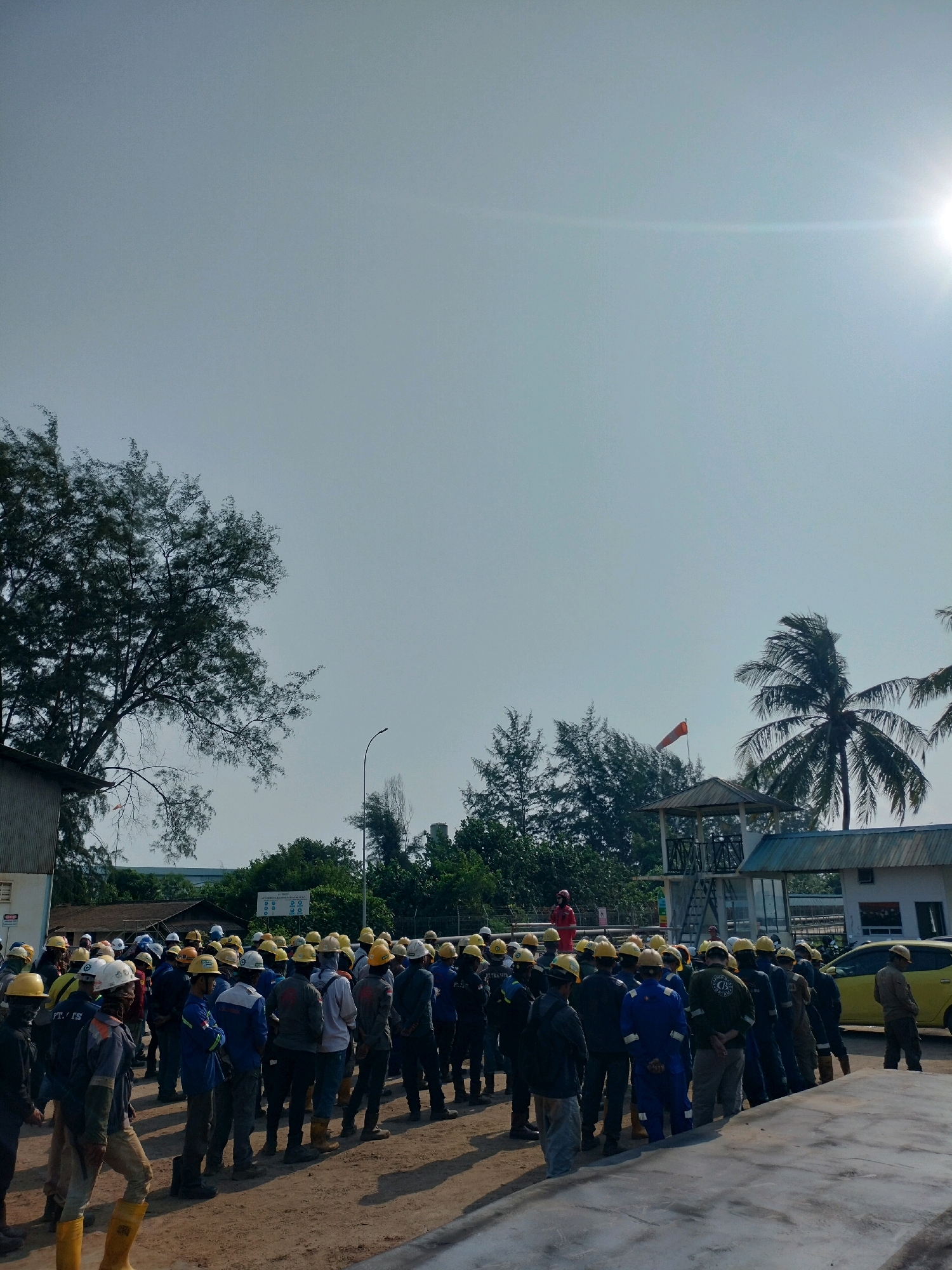 The image depicts a gathering of approximately 50 individuals, all wearing hard hats and safety vests, on what appears to be an industrial site in Batam, Indonesia. The individuals are standing in an open area, with some facing a person in a red jumpsuit standing on a platform in the background. The environment includes a building to the left, trees, a water tower with a windsock, and a yellow car to the right. The sky is clear and bright, suggesting it is daytime. The overall scene suggests a pre-work briefing or a safety meeting.