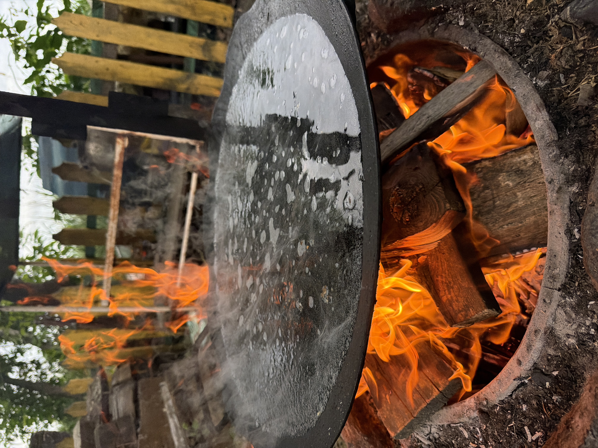 A wide, flat, black cooking surface, likely a griddle, is positioned over a fire pit. The griddle is filled with water that is actively boiling, evidenced by the steam rising and the bubbling surface. The fire beneath the griddle burns brightly with visible orange flames and glowing embers, consuming several pieces of firewood. The fire pit itself is a circular structure made of concrete or stone. In the background, a rustic wooden fence with yellow-painted pickets and some wooden structures are visible, suggesting an outdoor, possibly rural or backyard setting. Trees and greenery are also present in the background, partially obscured by the structures and steam. The overall scene depicts food preparation or cooking outdoors over an open flame.