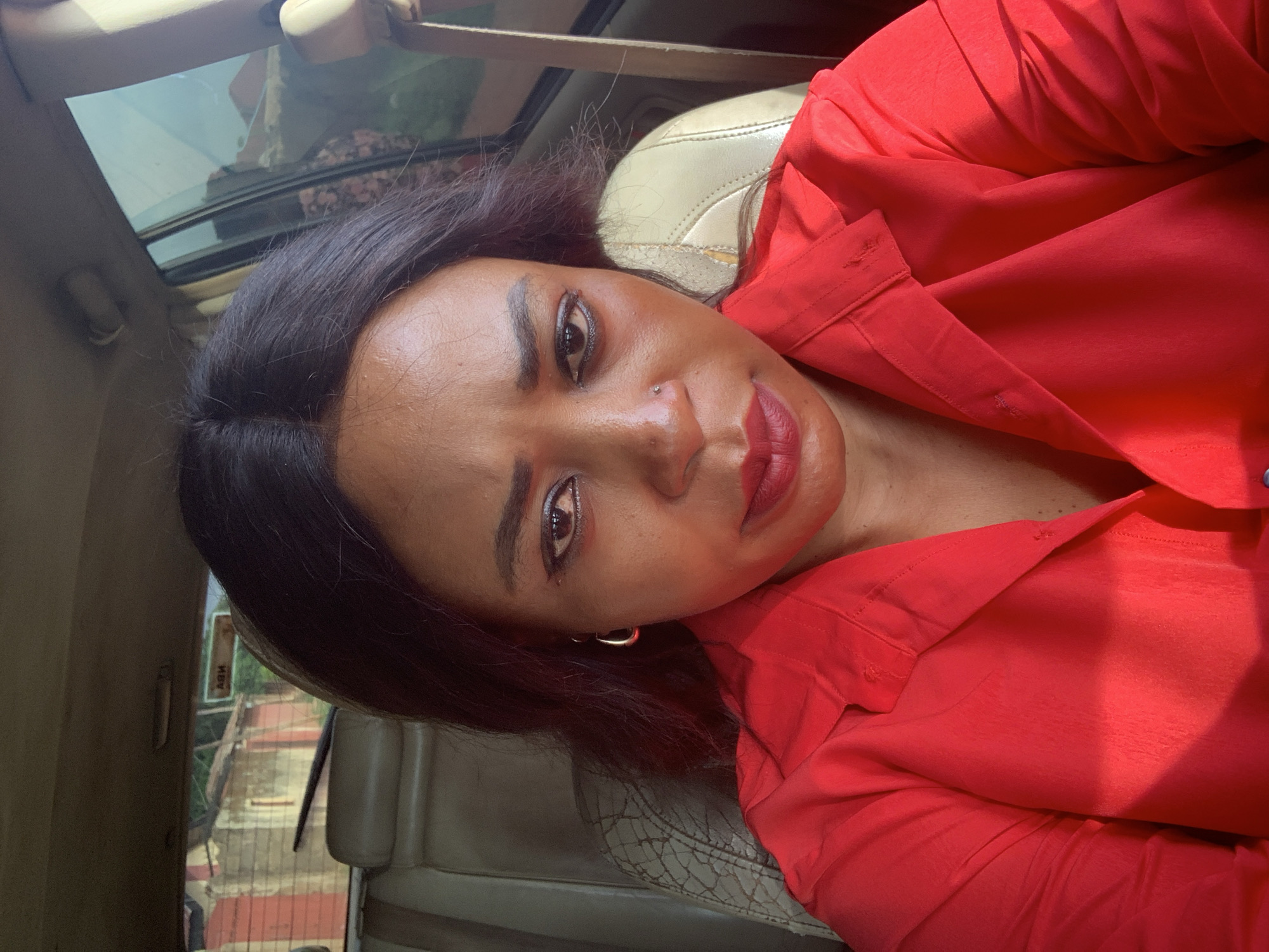 A woman with dark hair and dark eyes, wearing a red collared shirt, is seated inside a vehicle. She has hoop earrings in her left ear and a nose stud. Her face is turned towards the camera, and she is looking directly at it. The interior of the car, with its beige seats and headrests, is visible behind her. Sunlight filters into the car through the windows, casting shadows on her and the car's interior. Outside the car, through the windows, buildings with red and white striped walls and a blue sky are visible, suggesting an urban or suburban environment, possibly in Sabon Tasha, Nigeria.
