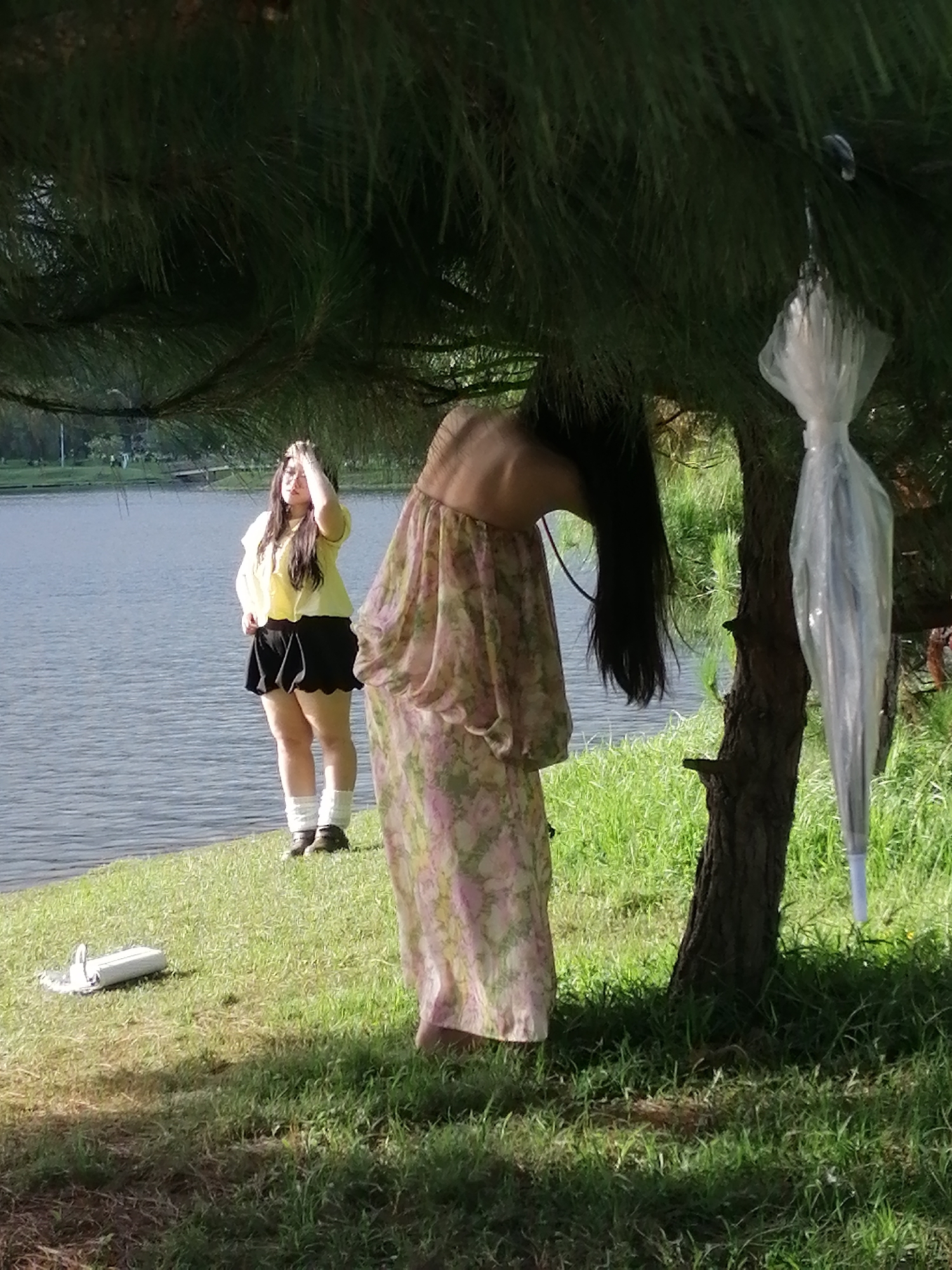The image depicts a park-like setting near a body of water, likely a lake, in Da Lat, Vietnam. In the foreground, a person is partially visible, wearing a floral dress and with their head bent forward, obscuring their face. Their long dark hair hangs downwards. To the left of this figure, a second person stands by the water's edge, looking to their right and shielding their eyes with their hand. This individual is wearing a yellow top and a black skirt, with white socks pulled up to their calves.
A tree with dark bark stands to the right of the frame, with pine needles from an overhanging tree creating a canopy. An umbrella, wrapped in clear plastic, is hanging from a branch of this tree. On the grassy bank in the foreground, a white object, possibly a water bottle or cooler, lies on its side. The ground is covered in green grass, and the water appears calm with ripples on its surface. The overall scene suggests an outdoor leisure activity in a natural environment.
