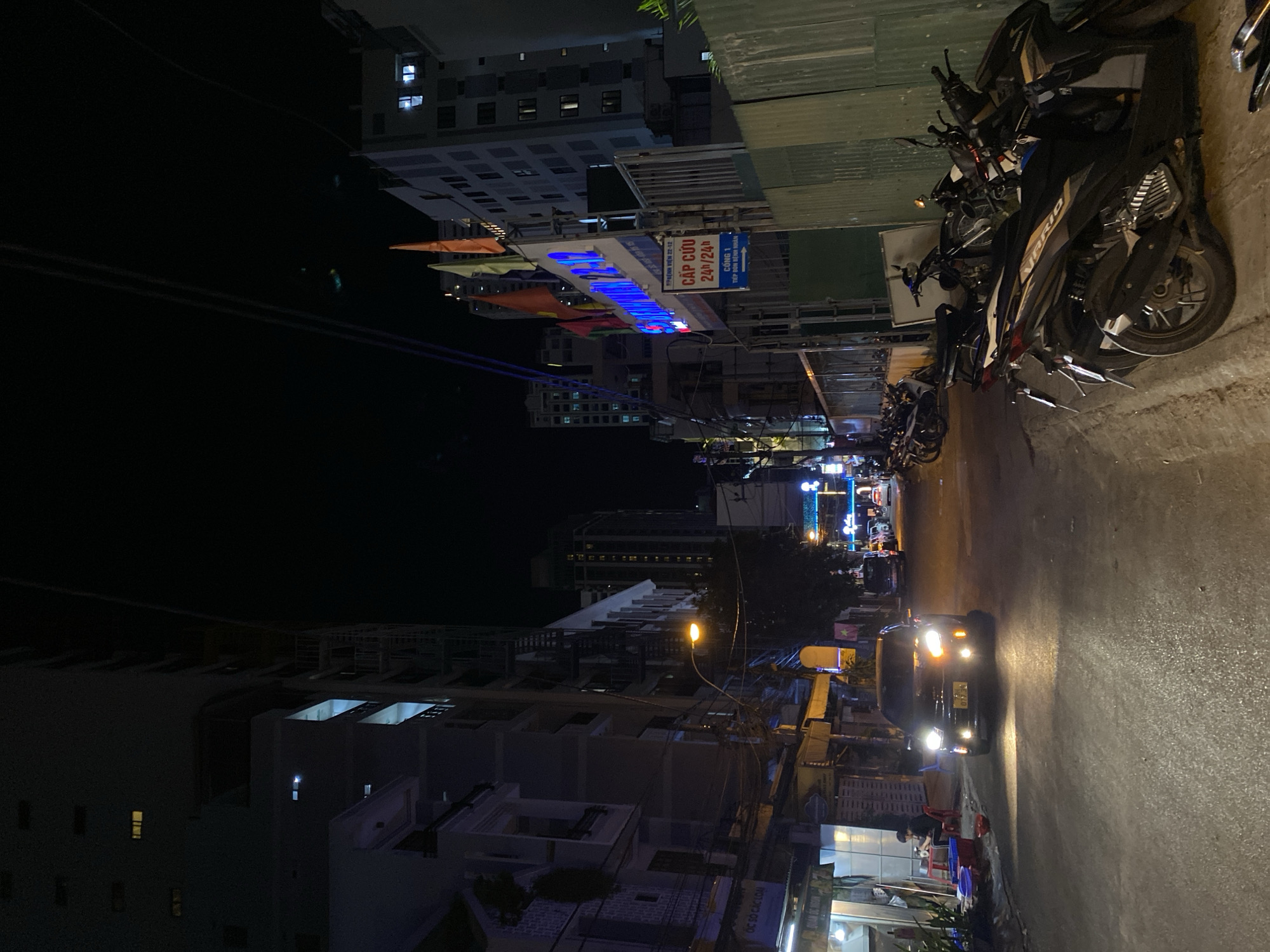 This is a nighttime, street-level, medium shot taken on a paved road in Nha Trang Ward, Vietnam. The scene depicts a moderately lit urban environment with several buildings lining both sides of the street. On the left, buildings appear to be of varying heights, some with lit windows and balconies. A street lamp casts a warm glow onto the road. A dark-colored car is the primary subject in the center of the frame, its headlights illuminating the asphalt. The car's license plate is partially visible. To the right of the road, a row of motorcycles are parked. Further down the street, more buildings and streetlights are visible, suggesting a commercial or residential area. The sky is dark, indicating it is nighttime.