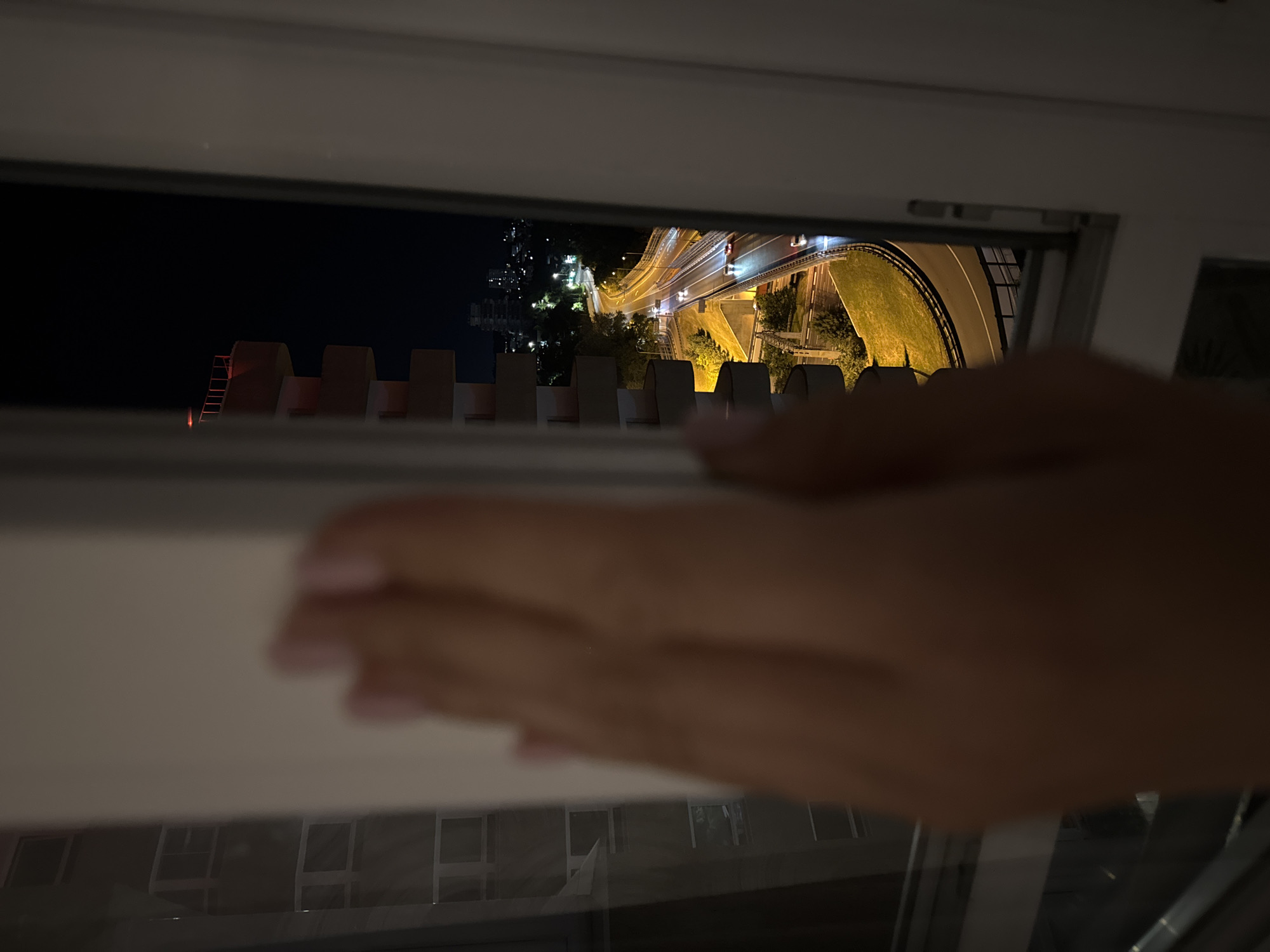 The image is a night time shot from an apartment in the city of Sochi, Russia, overlooking a highway interchange. A hand, partially blurred, is visible in the foreground, suggesting the viewer is looking out from behind a window or doorway. The highway is illuminated by streetlights, and cars can be seen driving on it, their headlights and taillights creating streaks of light due to the long exposure. The surrounding area features greenery and buildings, with some structures in the distance appearing illuminated. The architecture of a building with balconies is also visible on the left side of the frame. The overall scene depicts a typical urban landscape at night.