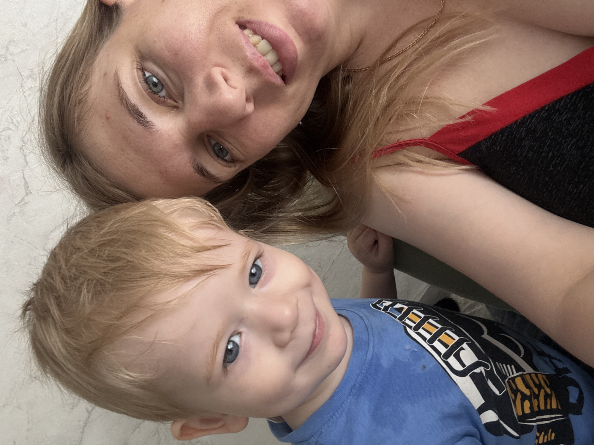 The media file is a close-up, selfie-style photograph of a woman and a young child, likely mother and son. They are positioned in a way that suggests they are lying down or leaning together, with the woman's face partially obscuring the top of the child's head.
The woman has blonde hair, light skin, and blue eyes. She is smiling widely, revealing her teeth, and appears to be looking towards the camera. She is wearing a red and black top. A delicate gold chain necklace is visible around her neck.
The child has blonde hair and blue eyes, and is looking directly at the camera with a gentle smile. He is wearing a blue t-shirt with a graphic design on it, featuring black, white, and yellow elements that appear to be part of a cartoon or toy vehicle. A small dark spot is visible on the fabric of his shirt.
The background is a light-colored, textured surface, possibly a wall. The lighting appears to be indoor, with no specific time of day or weather cues. The overall emotion conveyed is one of warmth, happiness, and affection between the two subjects. The location context provided is Kostroma, Russia. There is no visible text in the image.