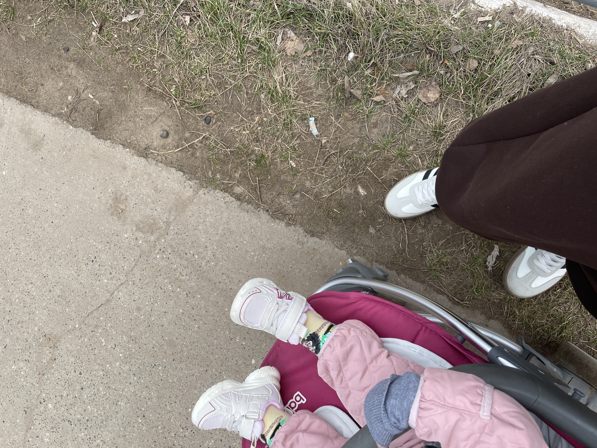 The image shows an overhead view of a person pushing a stroller along a sidewalk in what appears to be an urban area in Chaikovsky, Russia.
A child is in the stroller, wearing pink quilted outerwear, grey pants, and white sneakers. The adult pushing the stroller is wearing brown pants and white sneakers with black stripes. The stroller itself is a pink and white color, with a silver metal frame.
The surrounding environment consists of a concrete sidewalk on one side and an area of dirt and sparse grass on the other. There is some litter visible in the grassy area, including what looks like a discarded cigarette filter. The sidewalk appears cracked and worn, showing signs of use.