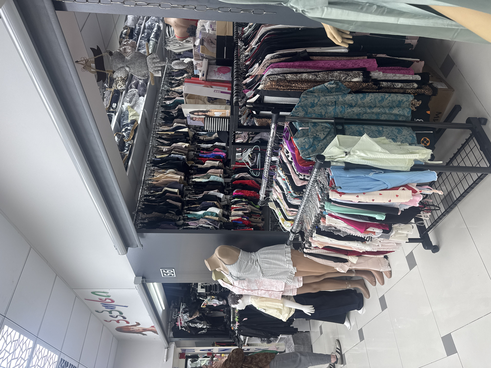 The image shows a clothing store with merchandise on display. The store is located in the city of Unknown, Unknown. Clothing is hung on racks and displayed on shelves. Mannequins are used to showcase clothing. A customer is visible in the lower portion of the image.