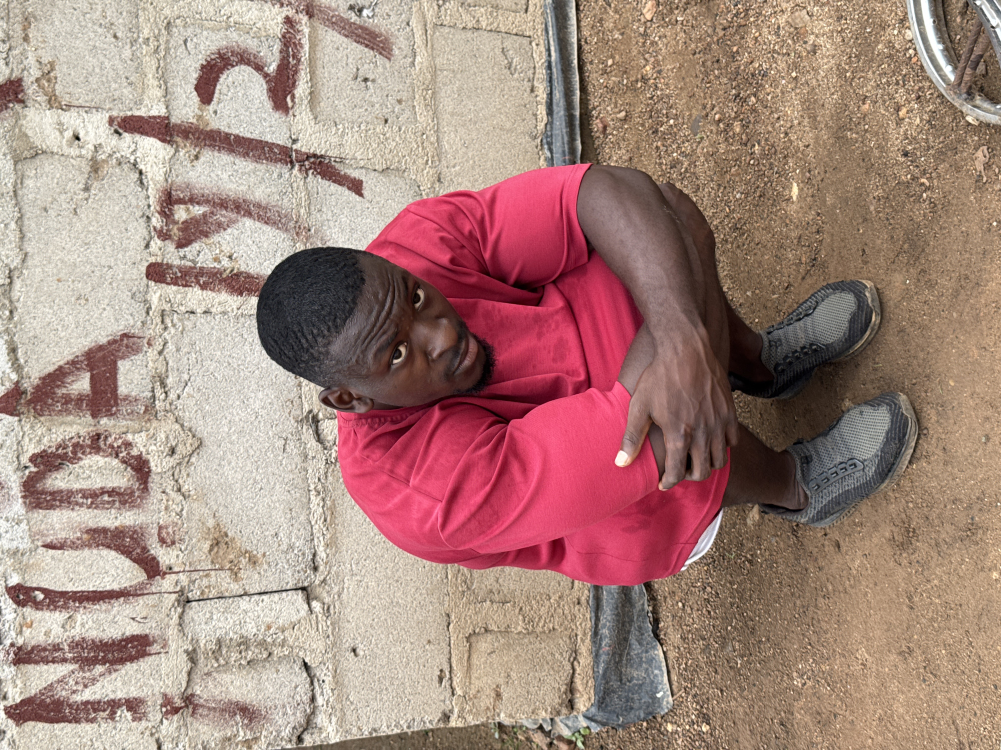 A Black male is squatting in front of a concrete wall with red graffiti. The graffiti on the wall reads "NUDA 19/2". The male is wearing a red t-shirt, white shorts, and grey sneakers. He is looking directly at the camera with his arms crossed. The ground in front of the wall is covered in dirt and gravel. A piece of metal, possibly from a bicycle wheel, is visible in the lower right corner of the frame. The scene appears to be outdoors, possibly in an alleyway or a neglected area of a city. The overall lighting suggests it is daytime.