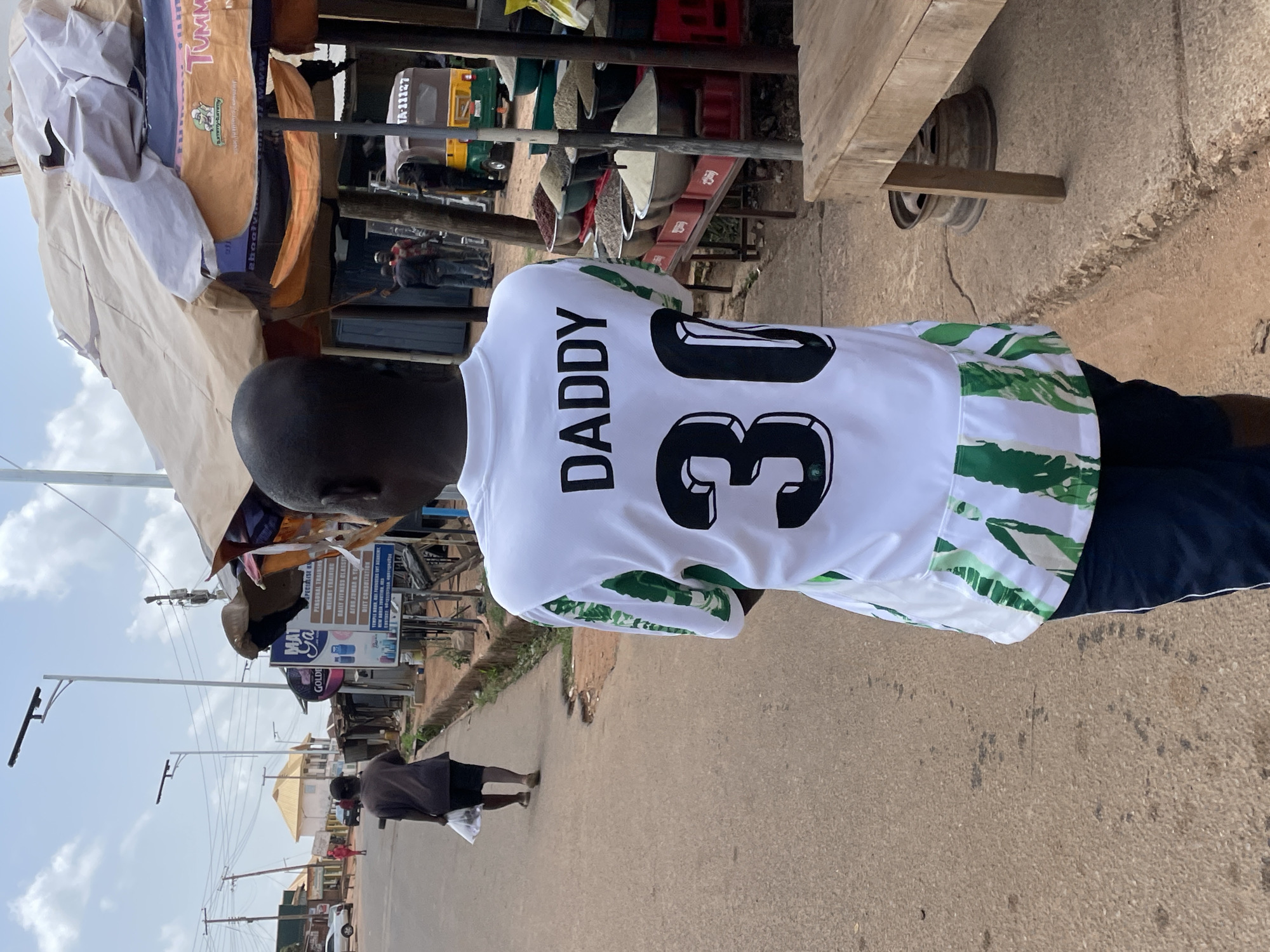 A person wearing a white jersey with "DADDY" and the number "30" printed on the back stands on a street. The jersey has green and white patterned sleeves. The person is facing away from the camera, and their head is shaved. Behind the person, there is a roadside shop with various items displayed. Across the street, there is a sign for "MAT Ga Specialist" with details about personal training, weight training, fitness classes, and diet consultation. Other people are walking in the background. The street is lined with utility poles and buildings. The environment appears to be an urban area, likely within the city of Jos, Nigeria. A yellow motor vehicle with registration "TA-11127" is near the shop.